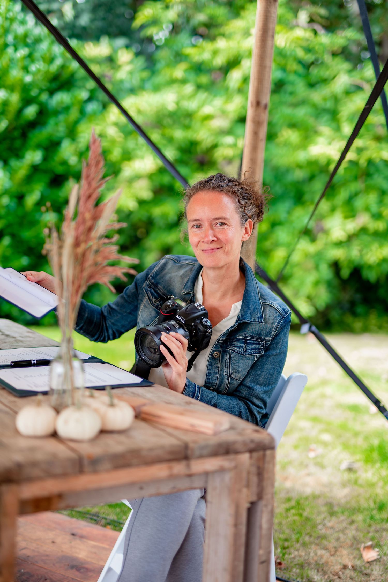 A photographer in a denim jacket sits at a rustic wooden table working with camera equipment in an outdoor setting.