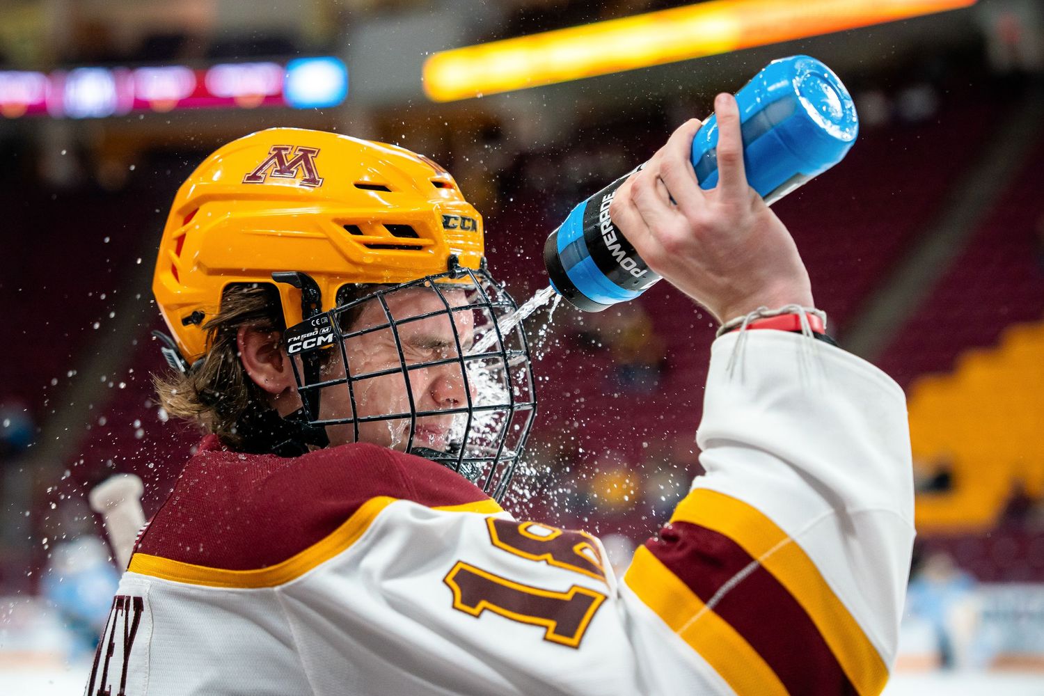 Minnesota Gophers hockey player in gold helmet squirts water bottle during break at arena.