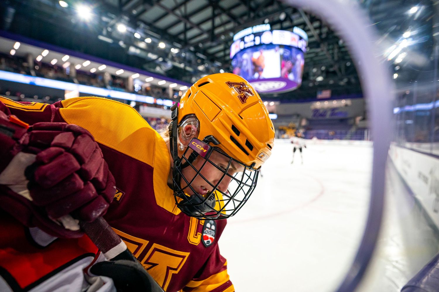 Hockey player in yellow and red uniform leans against the rink boards during a game in a large indoor arena.