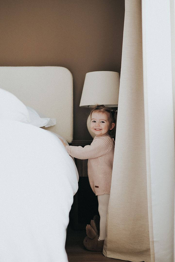 Smiling toddler in a cozy knit sweater stands beside a white bed and cream curtain next to a bedside lamp in a warm neutral bedroom.