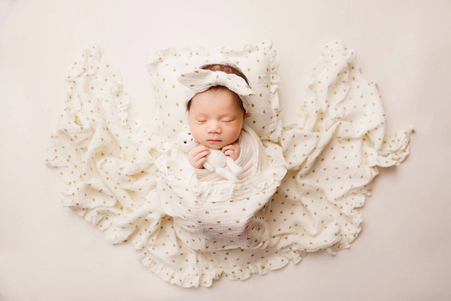 Sleeping newborn baby in a cream bow headband swaddled in a soft polka dot muslin wrap on a neutral background.