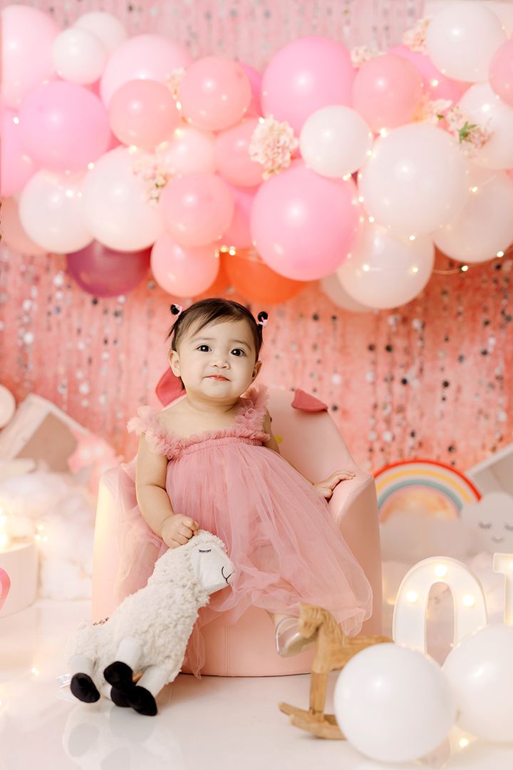 Baby girl in a pink tulle dress sits in a pink chair beside a plush lamb, with pink and white balloons and a sparkly backdrop behind her.