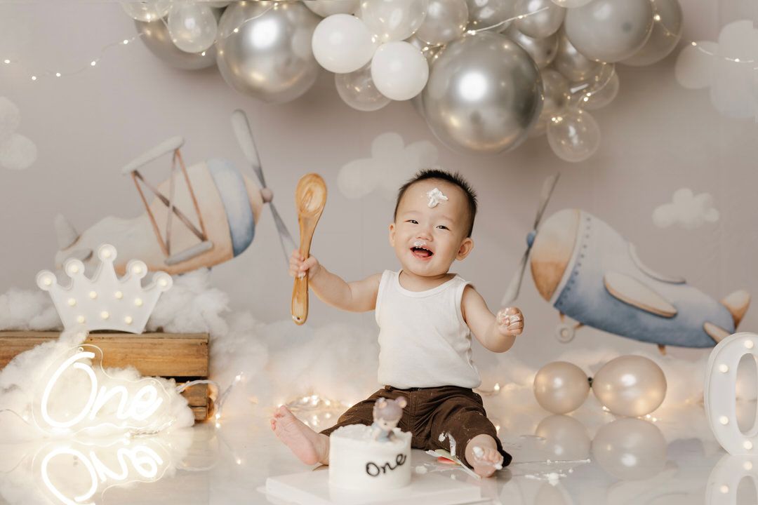 Smiling baby holding wooden spoons at a first birthday cake smash photoshoot with silver balloons, a crown, and a One neon sign.