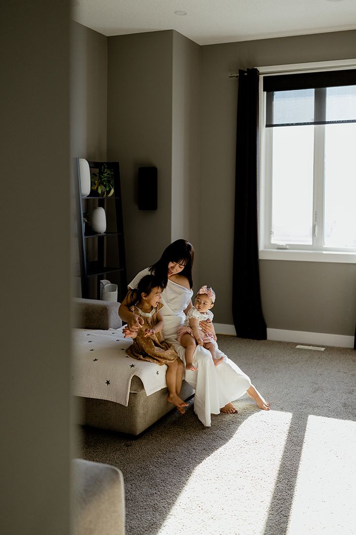 A mother in a white dress sits on a bed in a sunlit bedroom, cuddling two young children by a window.