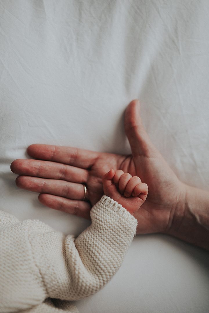 Tiny newborn hand in a cream knit sleeve resting on an adult's open palm against soft white bedding.