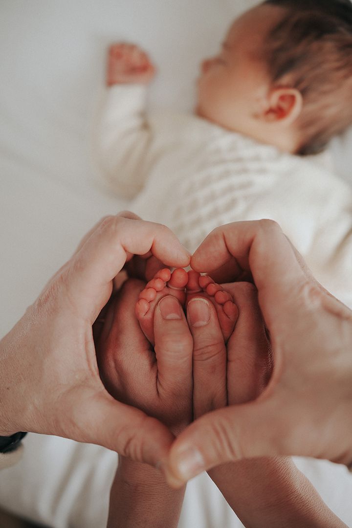 Adult hands form a heart shape around the tiny feet of a sleeping newborn baby in a cream knit outfit.