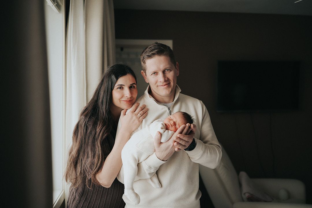 Smiling couple standing by a window in a cozy living room, gently cradling their sleeping newborn baby in a lifestyle family portrait.
