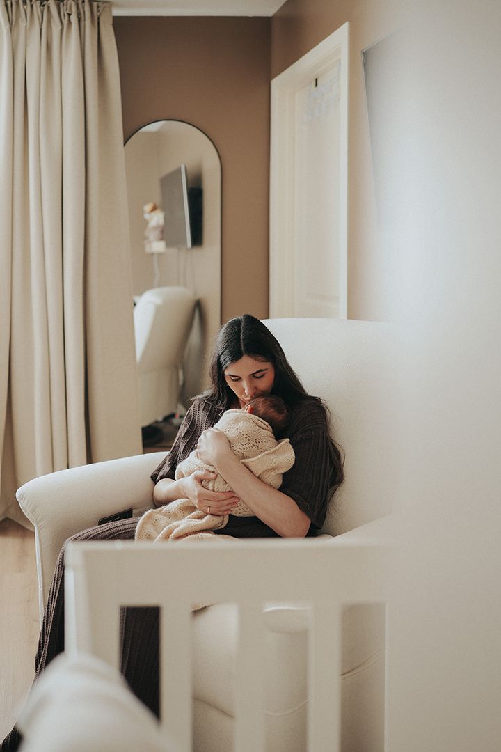Mother with long dark hair sits in a cozy white nursery chair, gently kissing her newborn baby wrapped in a soft knit blanket.