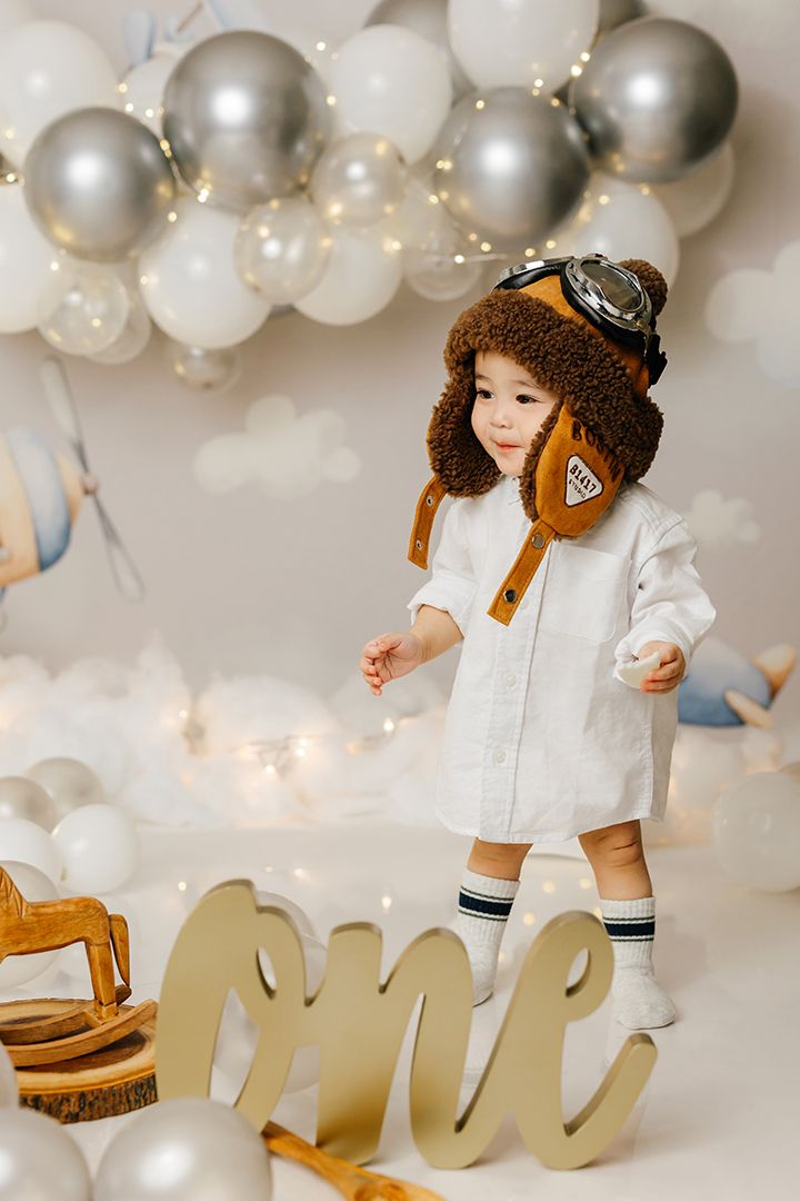 Toddler in a brown aviator hat and white shirt at a first birthday photoshoot with balloons, clouds and a gold ONE sign.