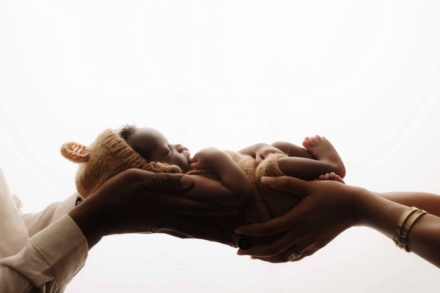 Two hands gently cradle a sleeping newborn baby wearing a knit bear hat against a soft, bright white background.