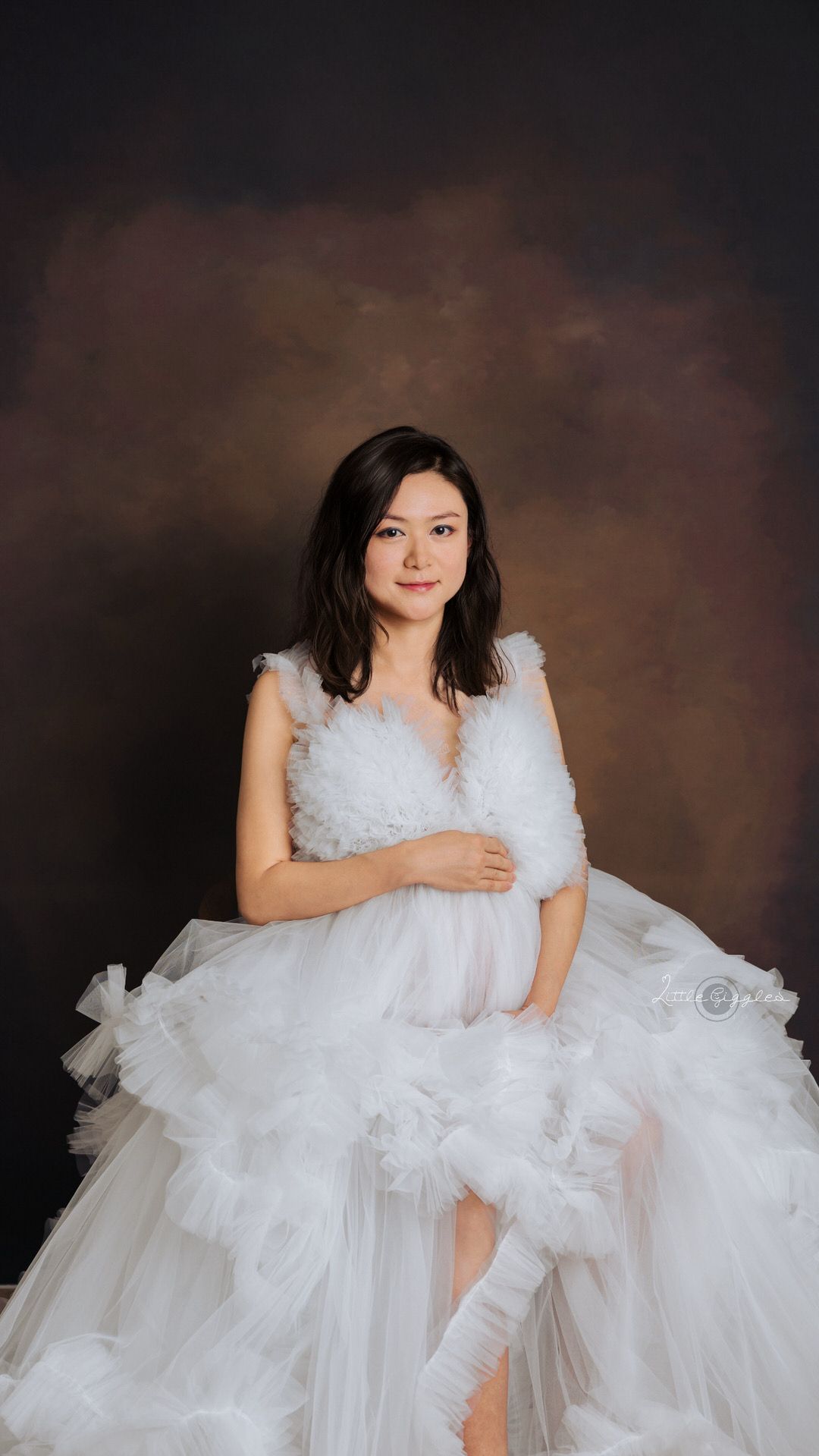 Woman in a flowing white maternity gown seated in a studio.