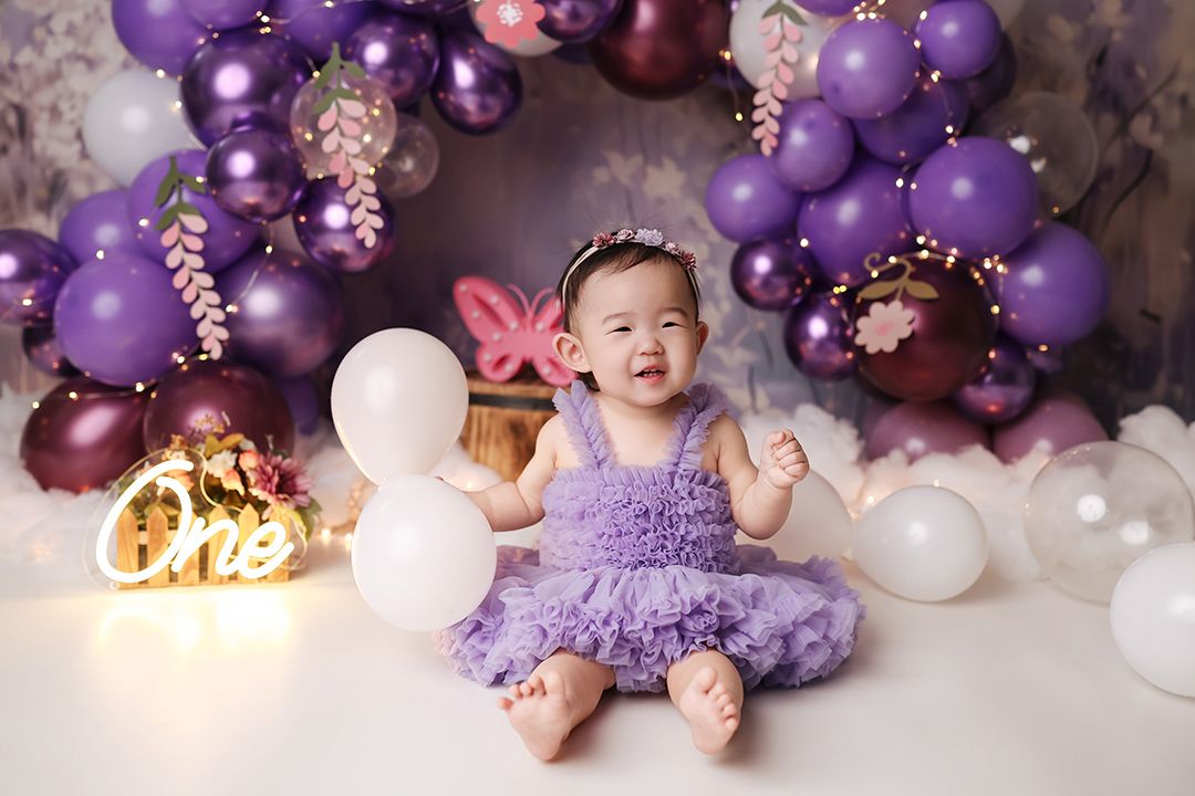 Baby girl in a purple tutu dress and headband sits smiling among purple and white balloons at a first birthday cake smash photoshoot.