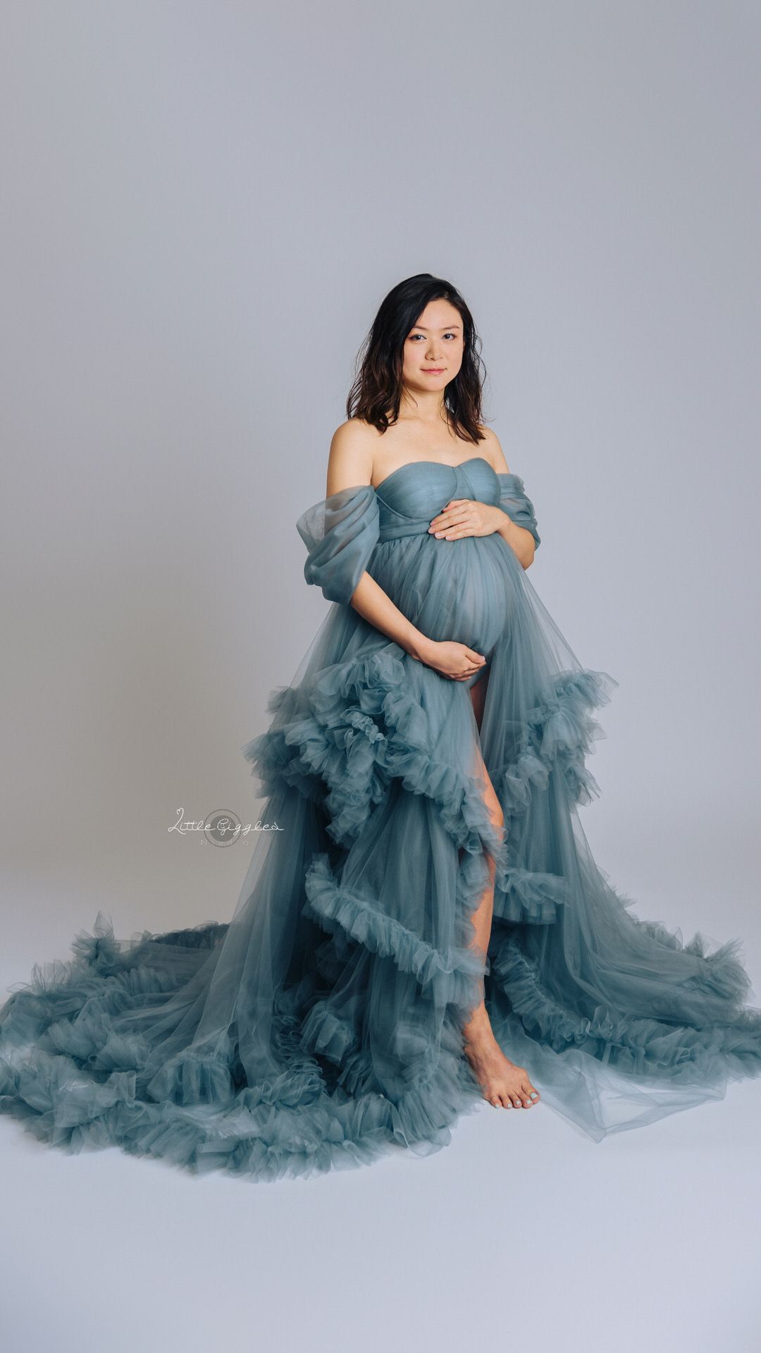 Pregnant woman in an off-the-shoulder blue-gray tulle maternity gown with ruffled train cradling her belly in a studio portrait.