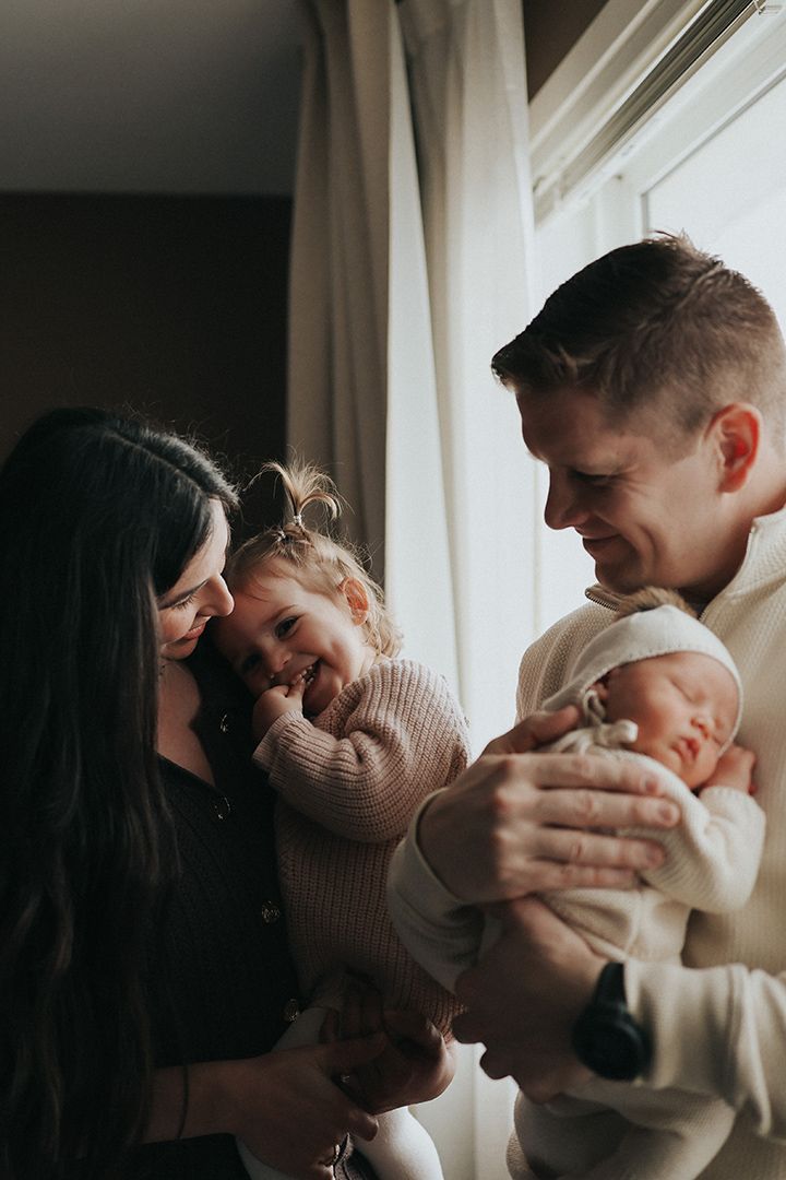 Smiling parents hold their toddler daughter and sleeping newborn baby in a cozy, softly lit family portrait by a window.
