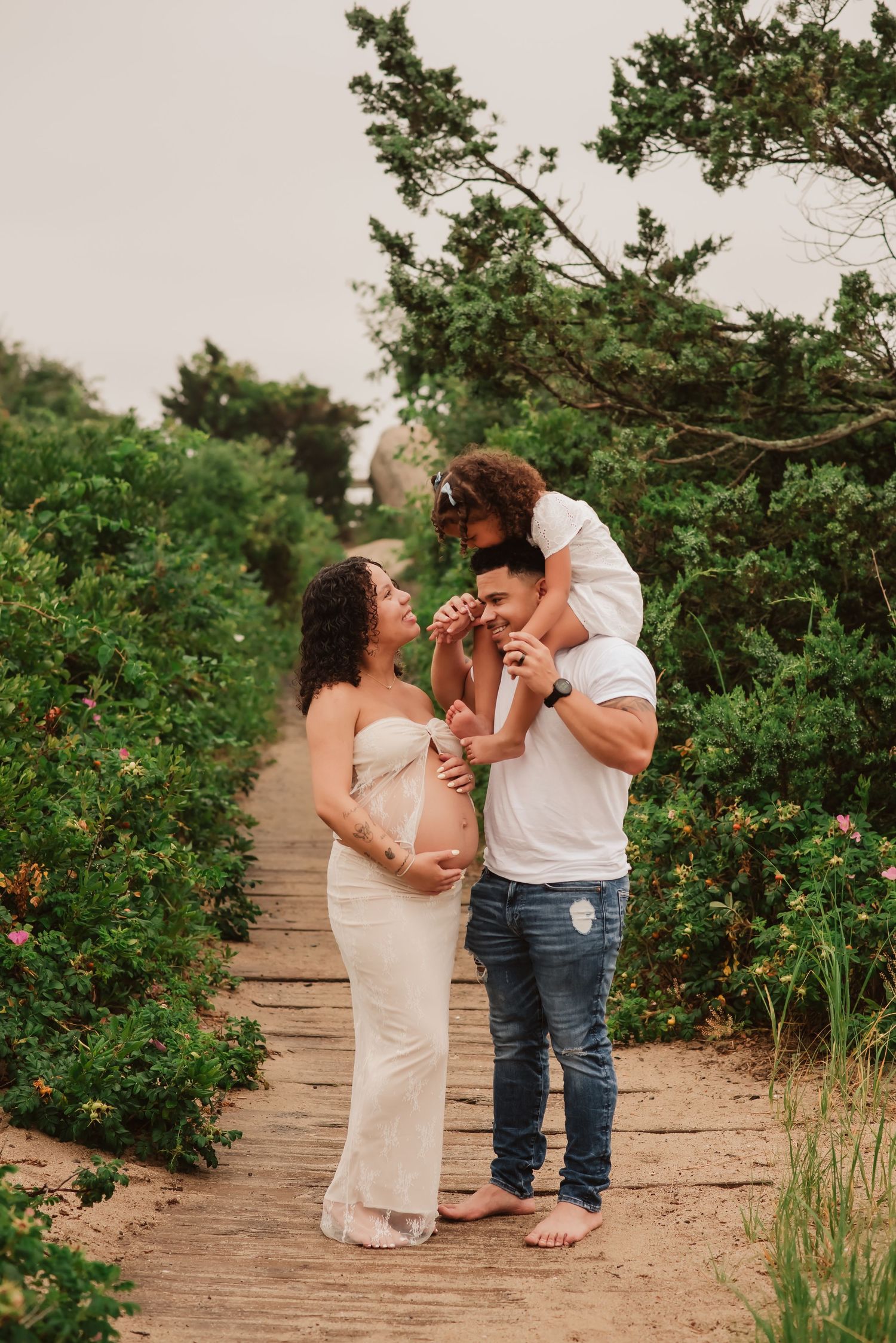 A family shares a tender moment on a wooden boardwalk surrounded by lush green coastal vegetation.