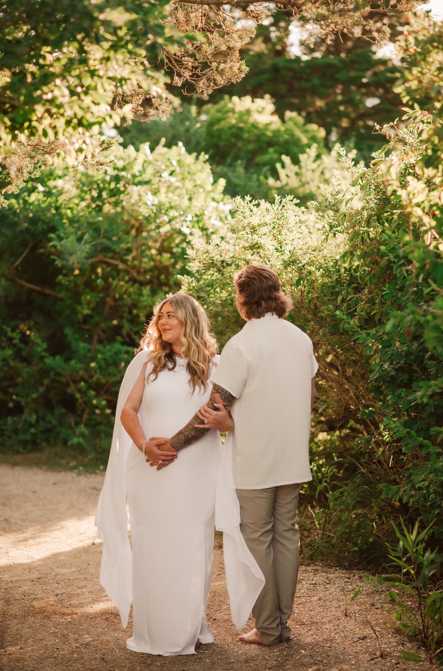 A couple in white clothing stands together on a sunlit garden path surrounded by blooming greenery and flowers.