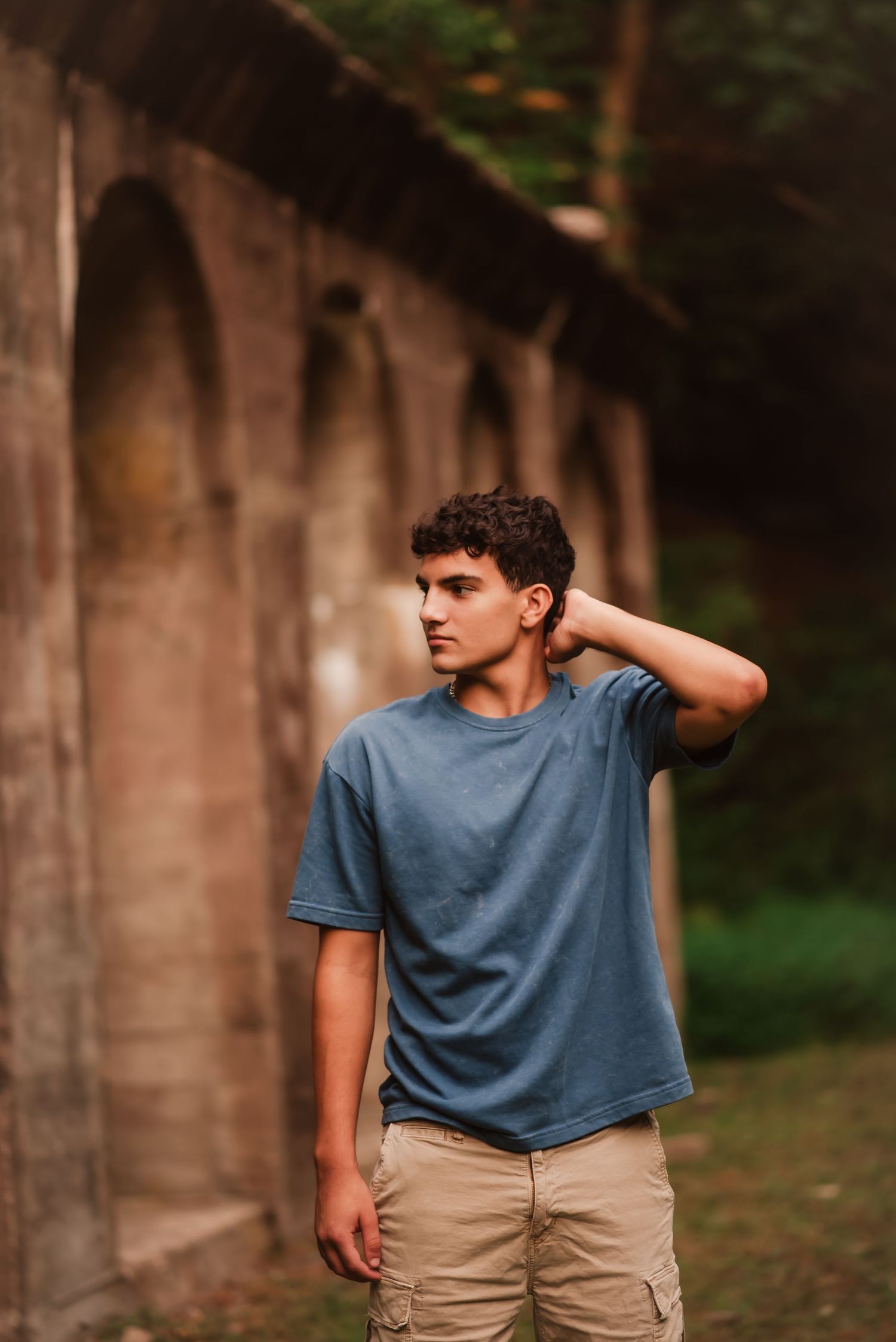 Young person in a blue t-shirt and khaki pants poses thoughtfully against a stone wall in an outdoor setting.