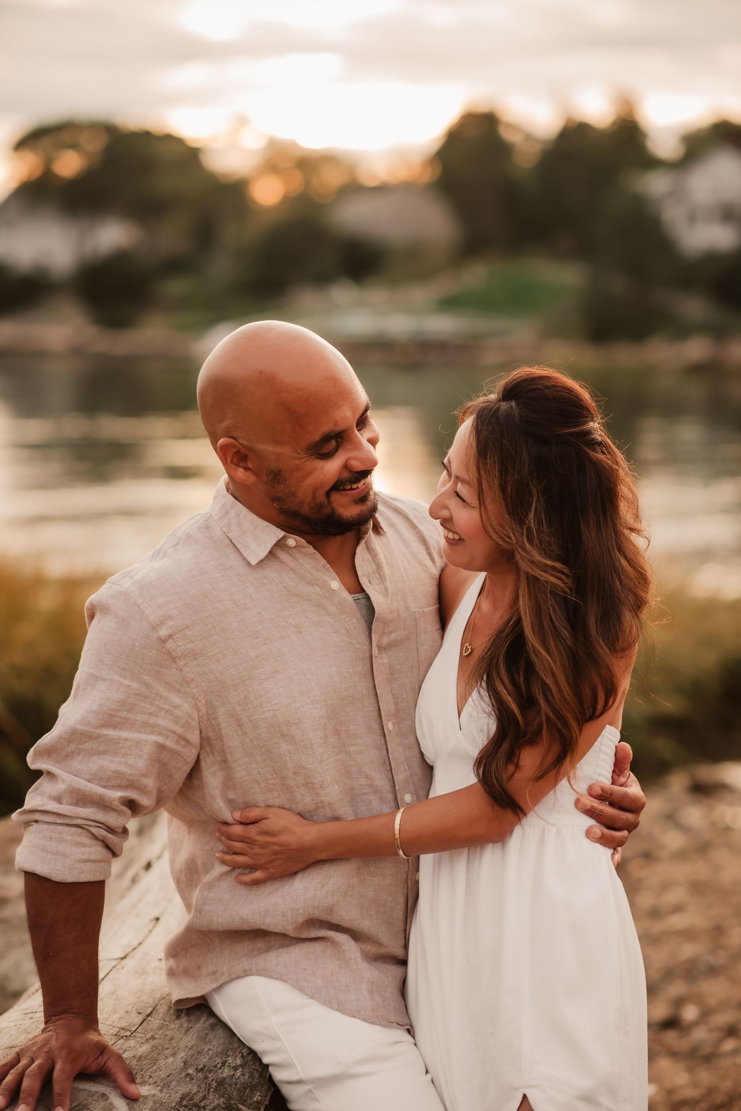 A couple shares an intimate moment at sunset by a lake, dressed in white and linen clothing.