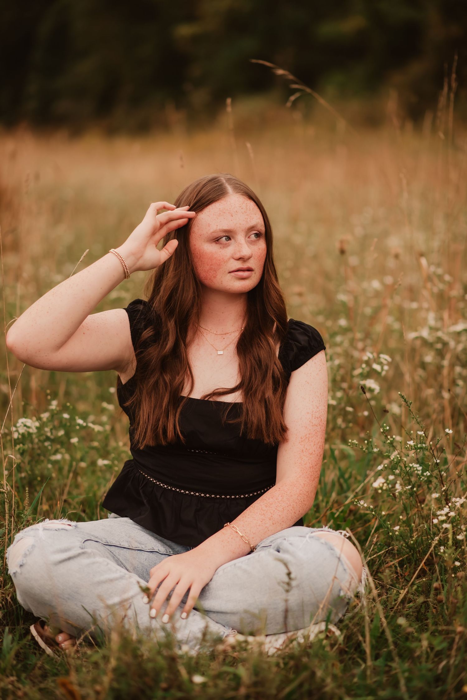A person with long auburn hair sits in a wildflower meadow wearing a black top and light colored pants at sunset.