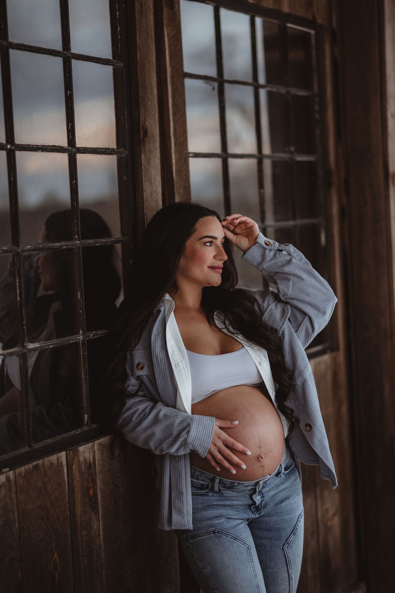 Pregnant woman in casual attire poses by rustic window during golden hour maternity photoshoot.