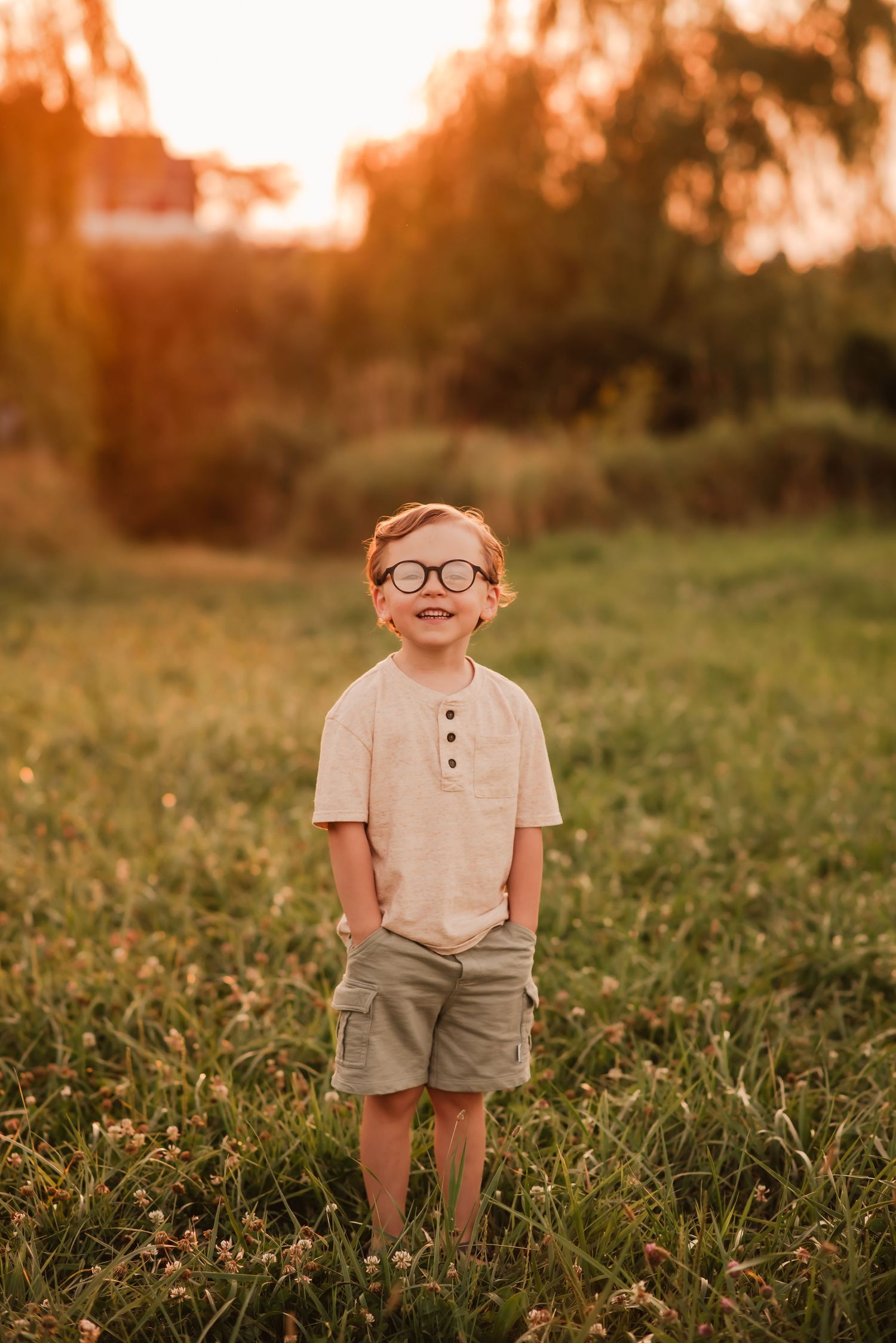A young child wearing glasses stands in a grassy field at sunset wearing a beige shirt and khaki shorts.