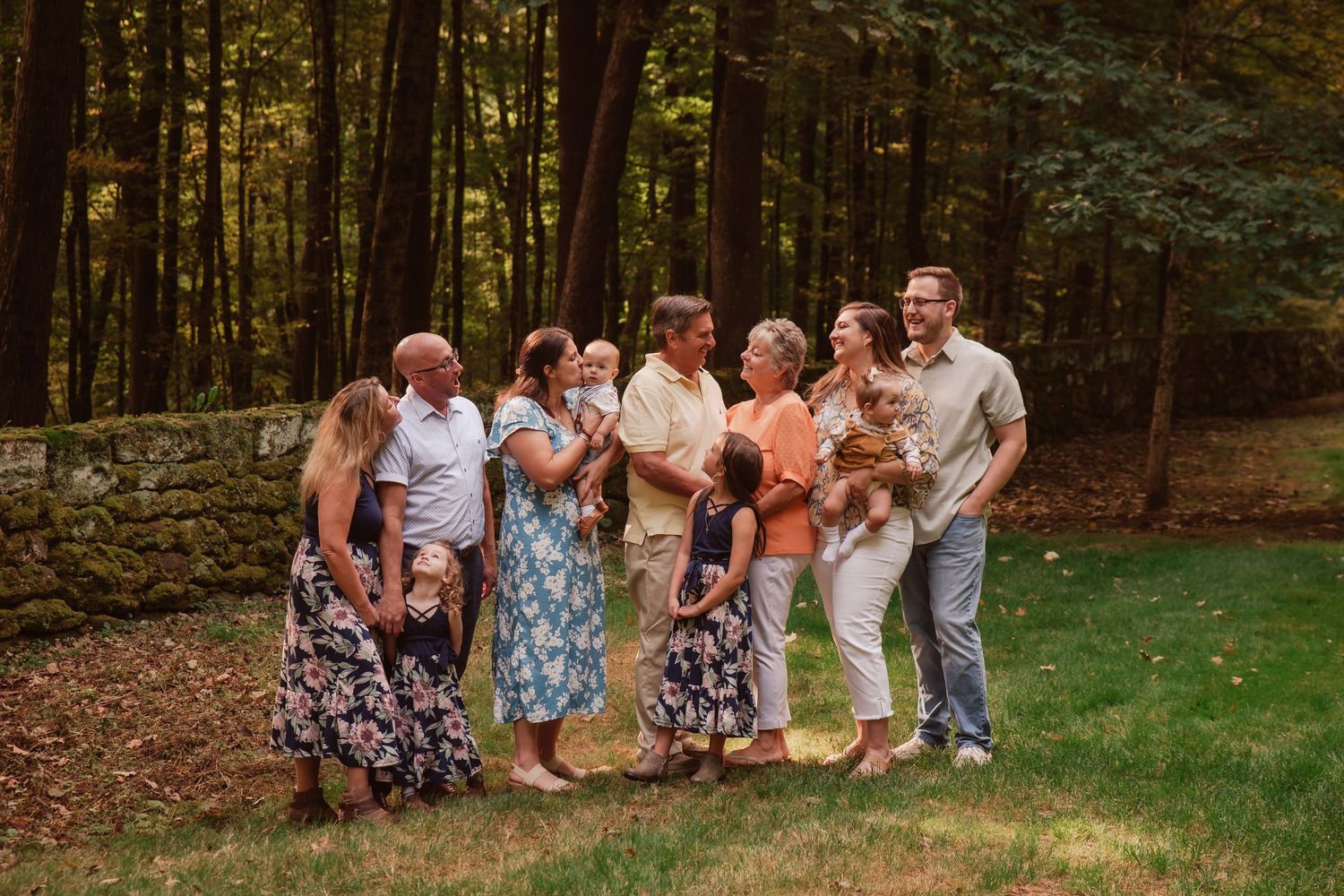 A group of people sit and laugh together on a grassy slope at the edge of a wooded area during a photo session.