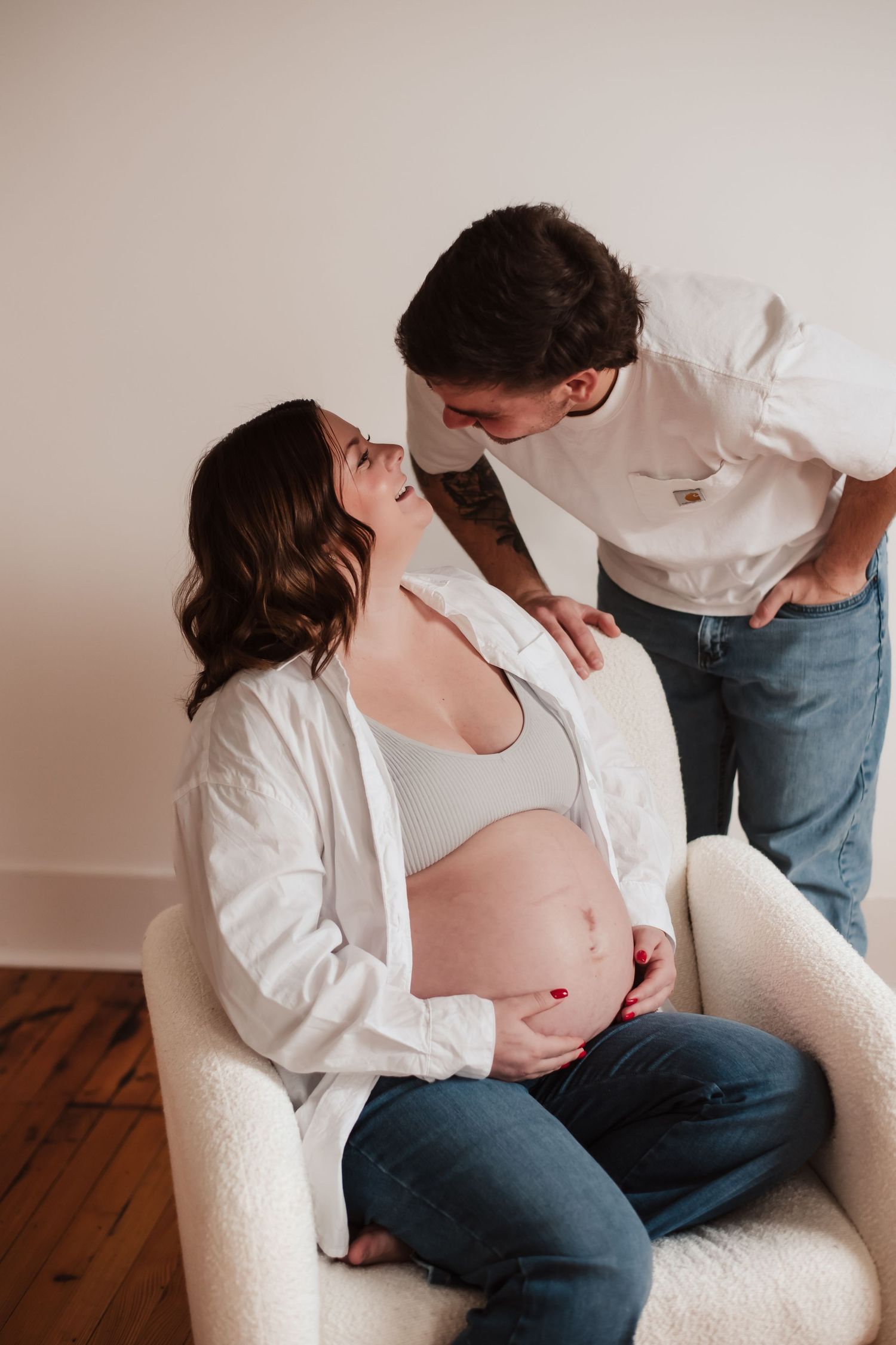 Intimate maternity photo of expecting couple sharing tender moment in white chair on hardwood floor.