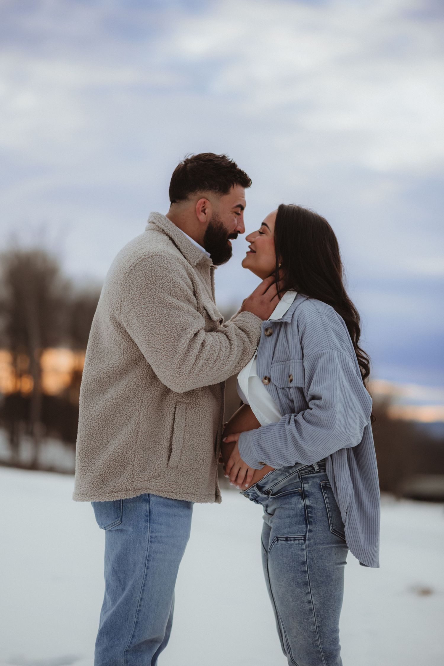 A couple in matching beige sweaters and jeans share tender moments in a snowy winter landscape at sunset.