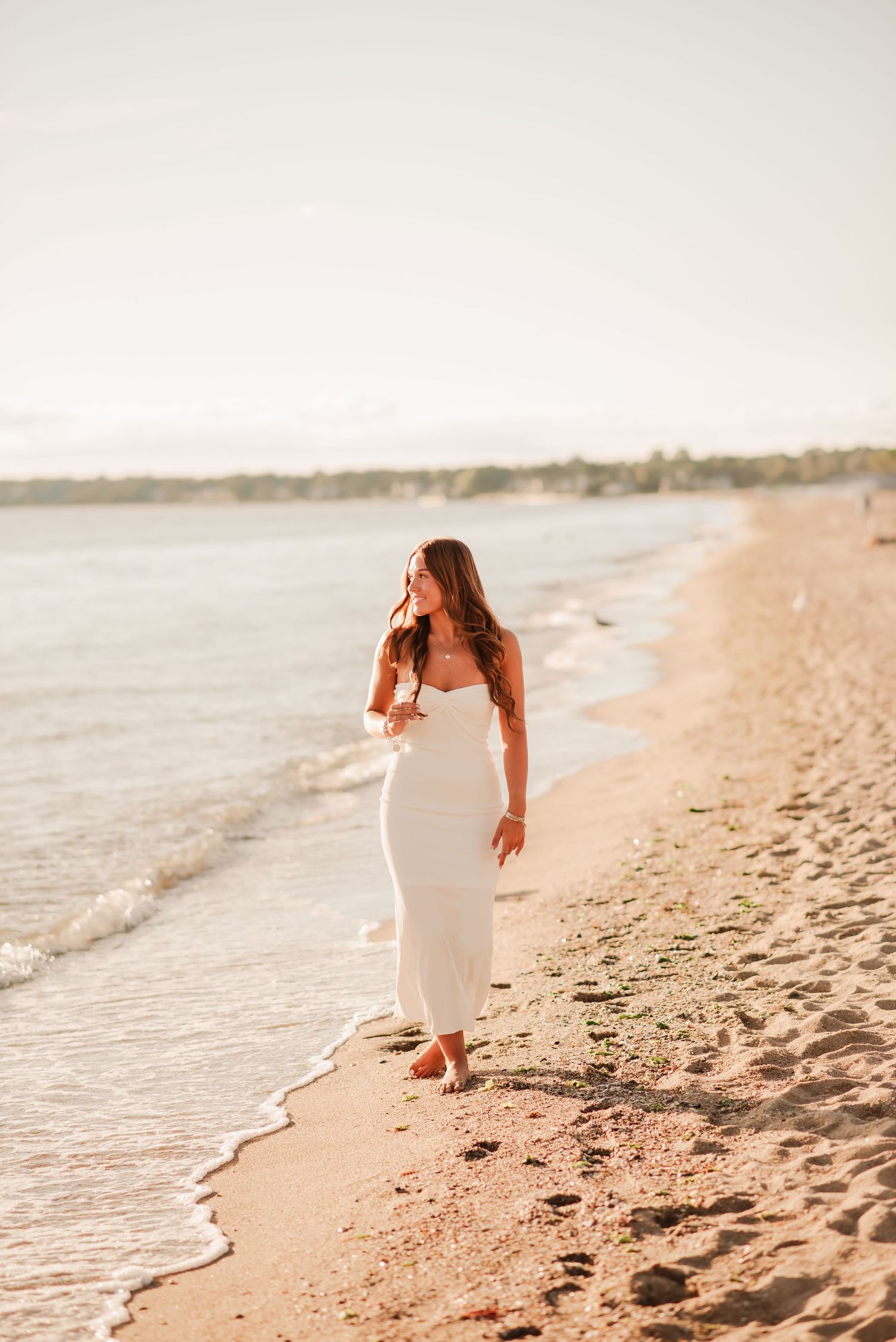 Woman in white dress walks along sandy beach at sunset with waves gently lapping at shore.