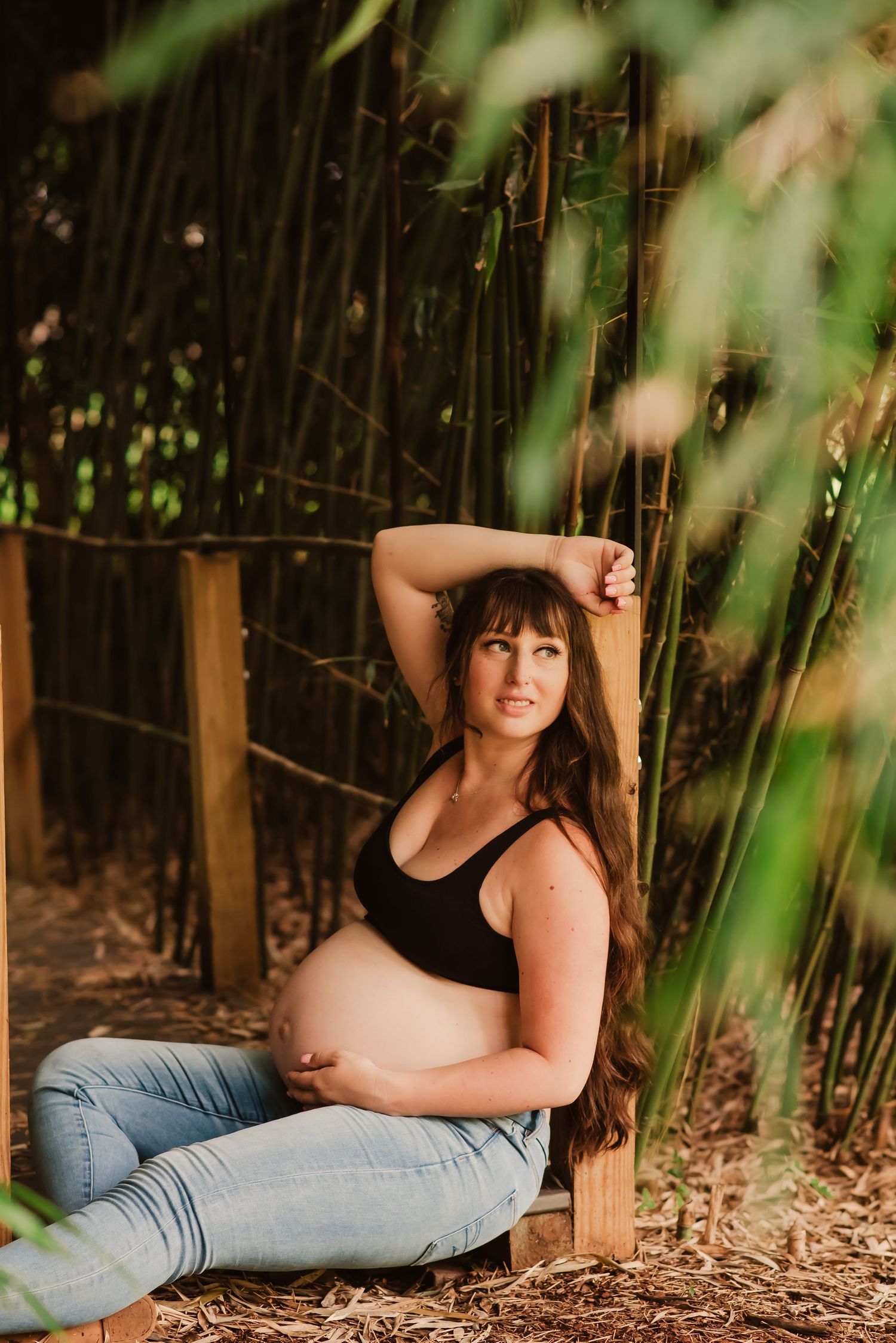 A person in jeans poses outdoors on a ground covered in leaves with bamboo trees in the background.