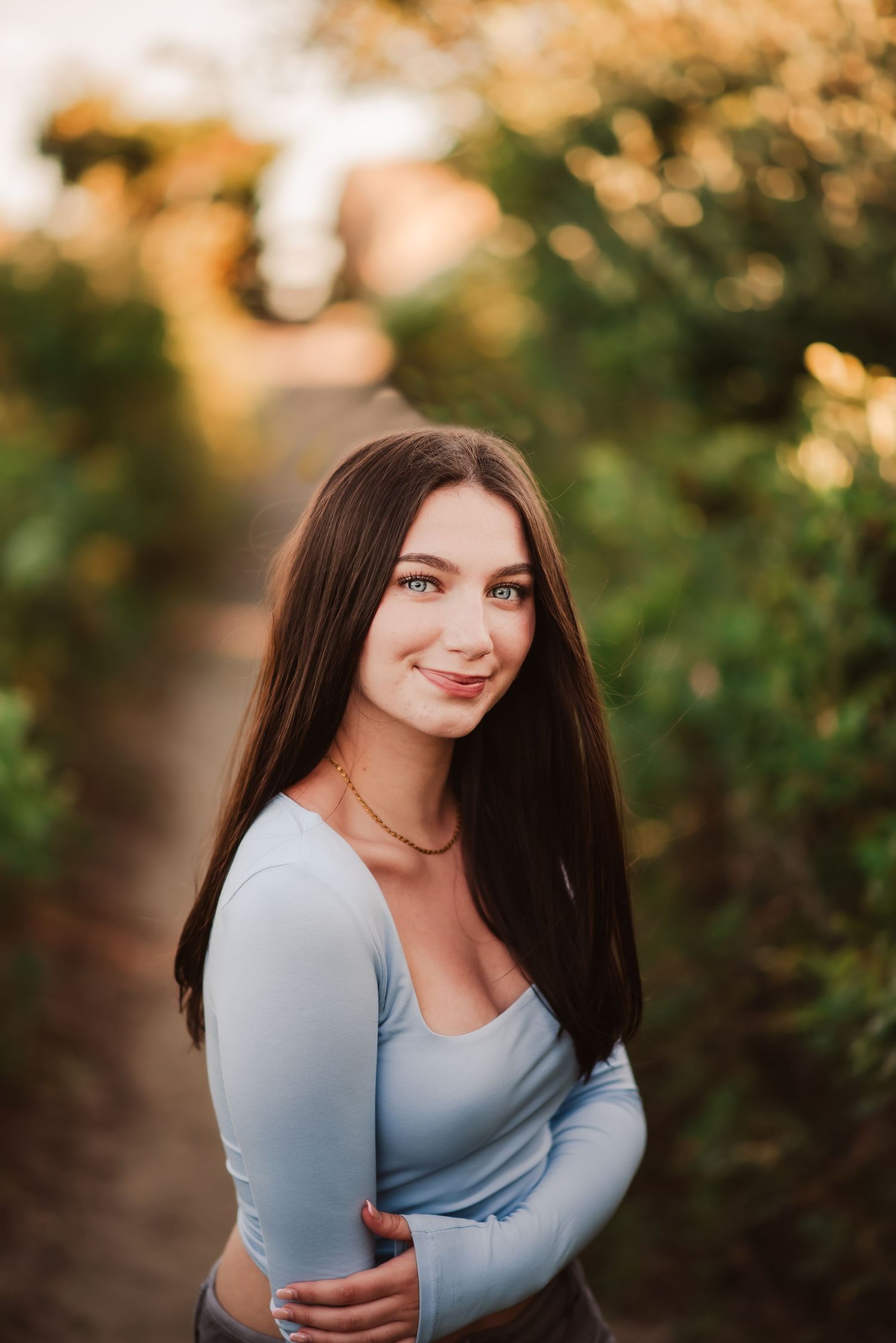 A person in a light blue long-sleeve top poses outdoors during golden hour with green foliage in the background.