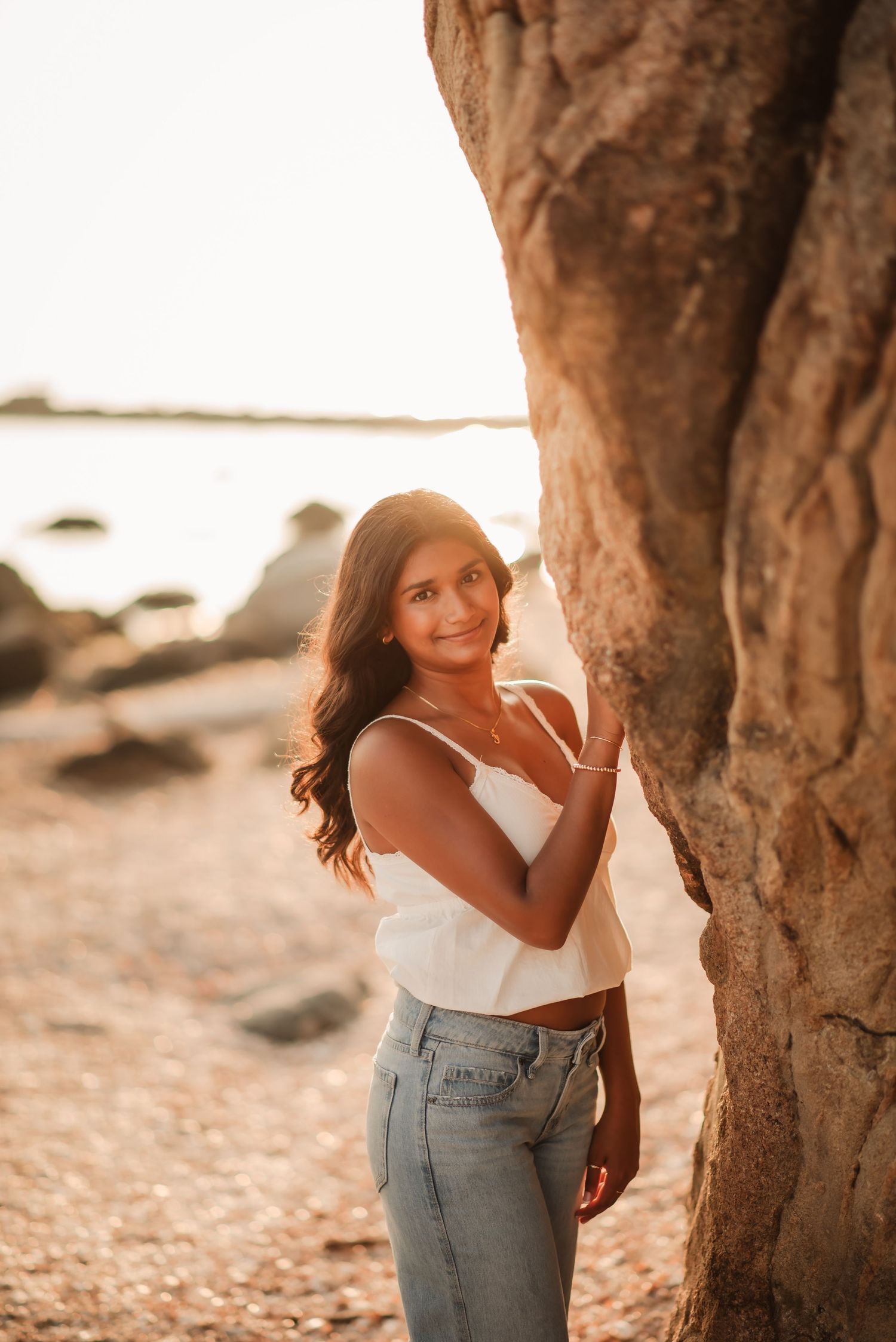 A person in a white top and jeans poses playfully among trees at sunset on a beach.