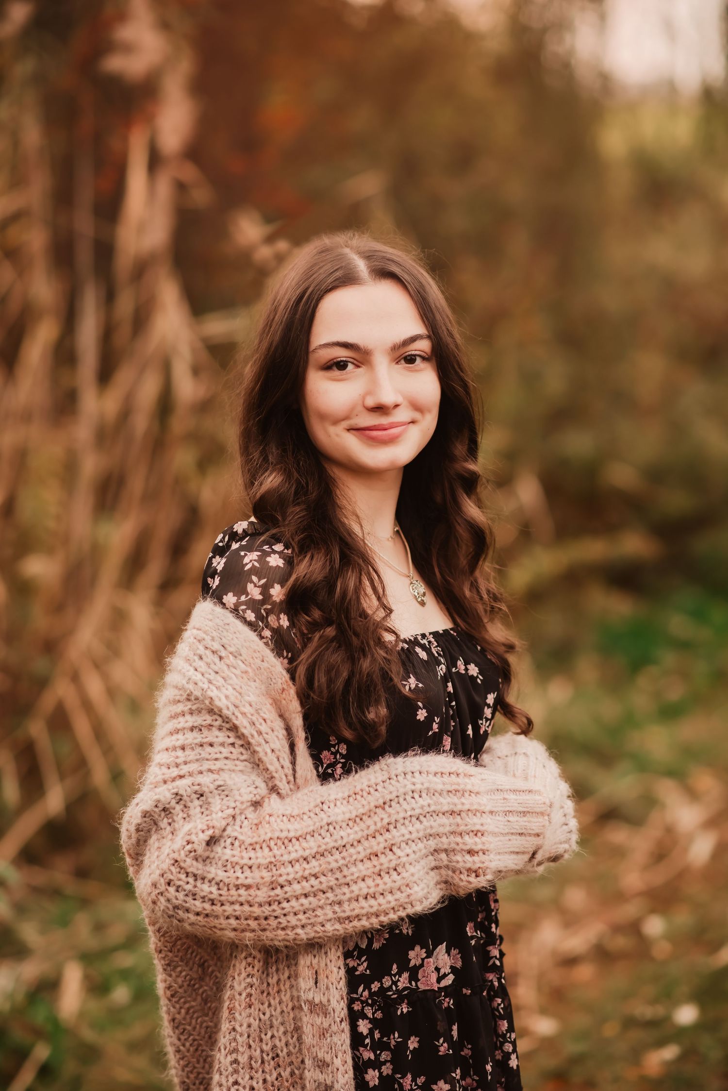 Young woman in floral dress and beige cardigan poses outdoors in autumn setting with natural lighting.