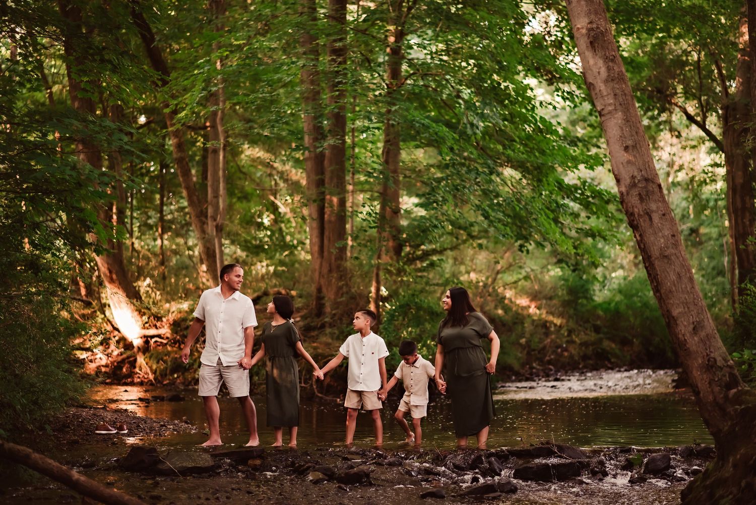 A family walks together through a sunlit forest path surrounded by tall trees and lush greenery.