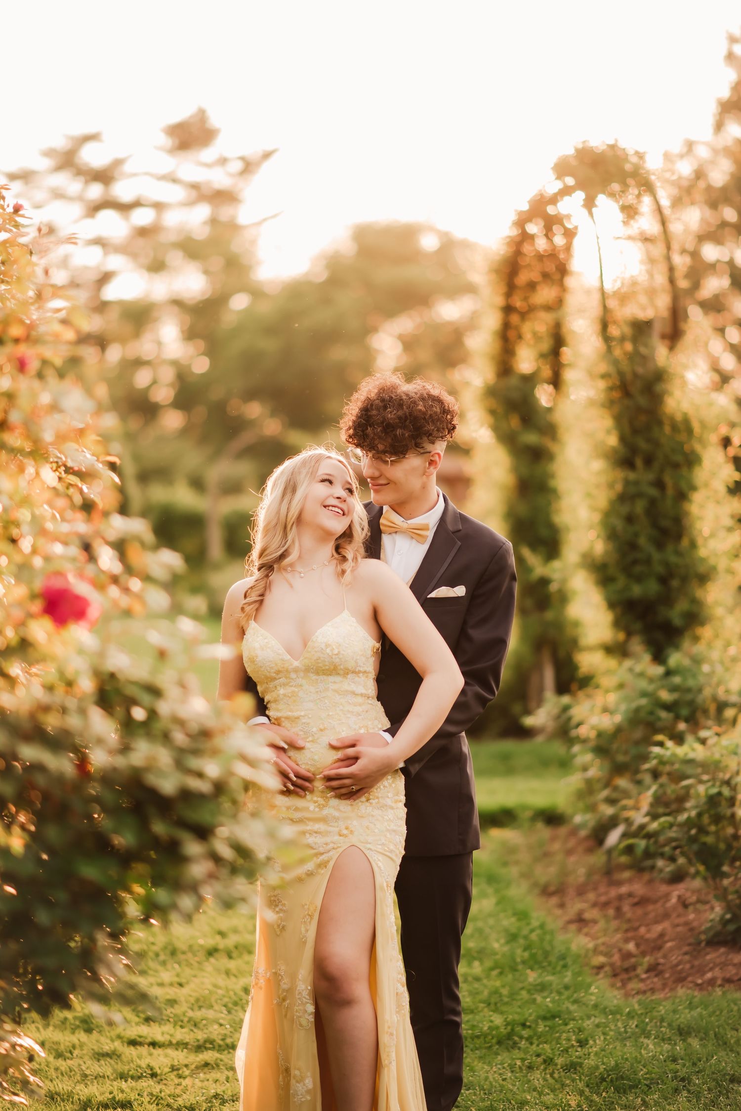 A romantic couple embraces in a sunlit garden during golden hour with roses and greenery in the background.