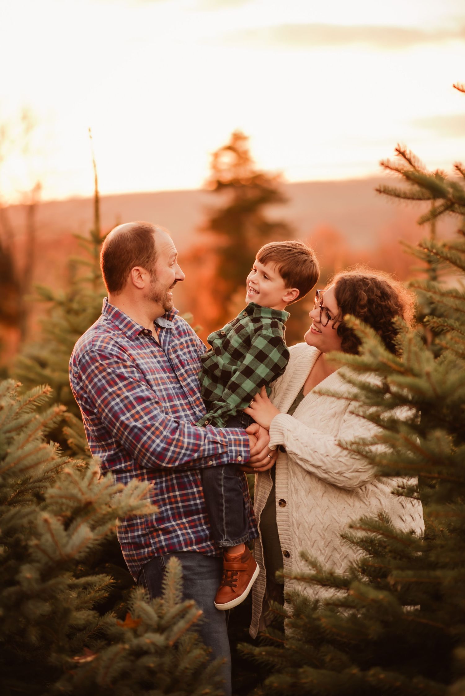A couple shares a playful moment among evergreen trees at sunset during a holiday photoshoot.