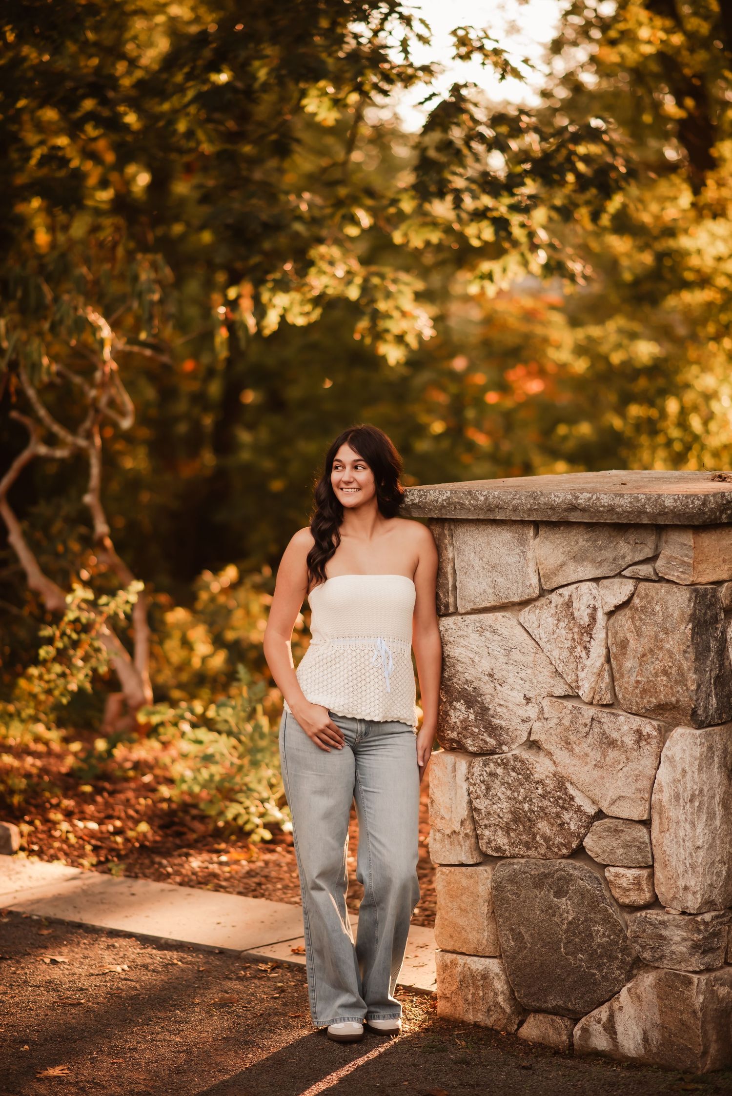 A person in a white top and jeans sits on a rustic stone wall surrounded by autumn foliage at golden hour.
