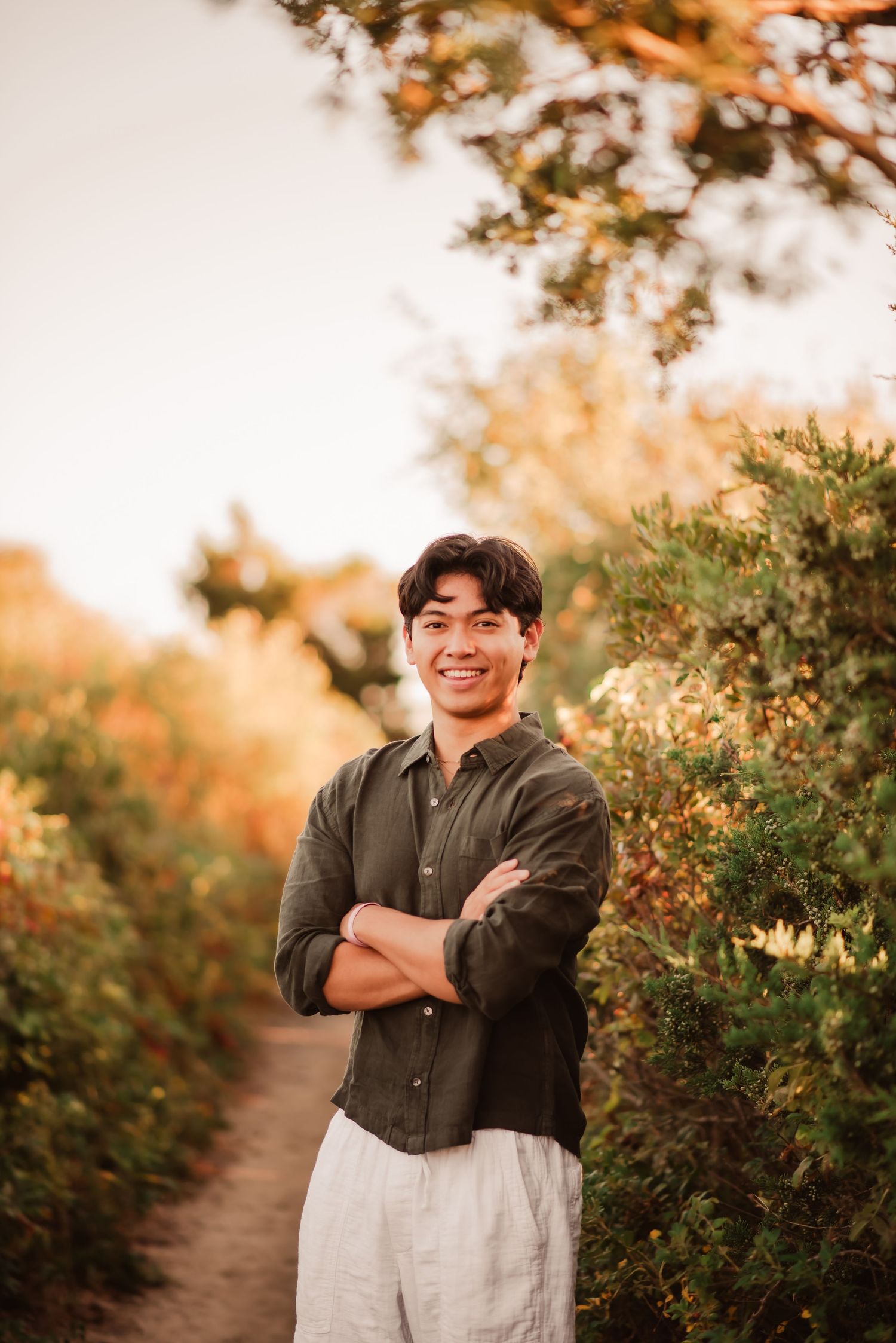 A person in a dark shirt and light pants stands smiling on a nature path during golden hour with autumn foliage.