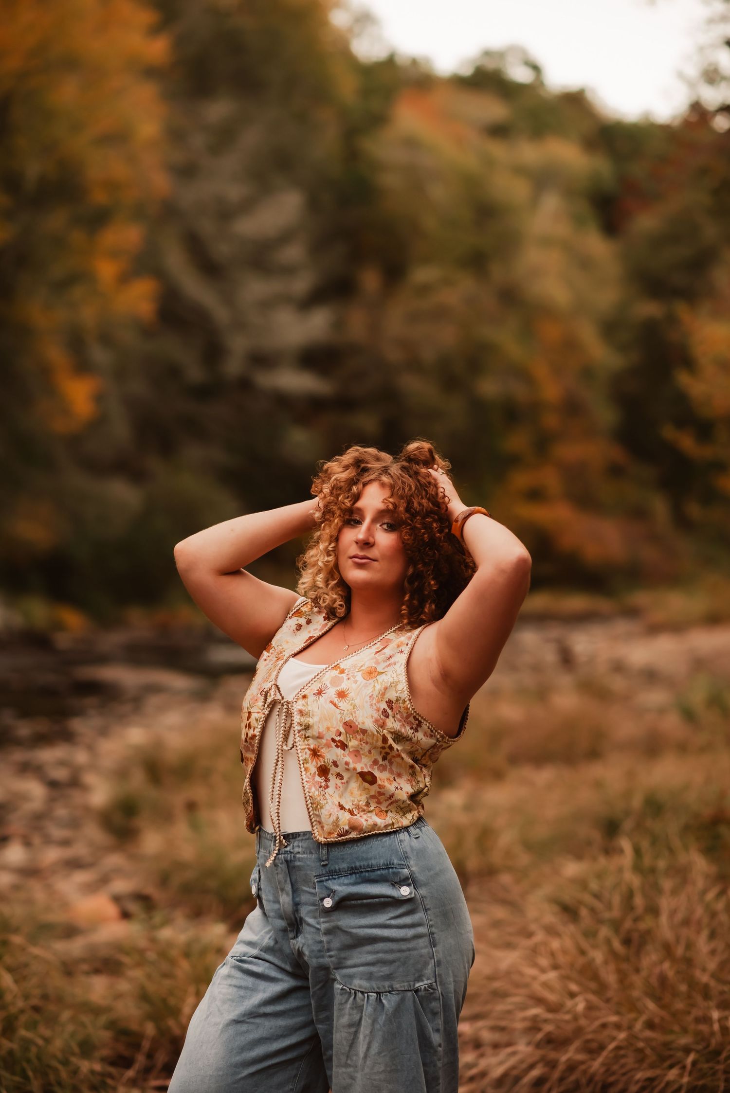 A person in denim overalls poses outdoors against an autumn backdrop with warm fall colors and foliage.