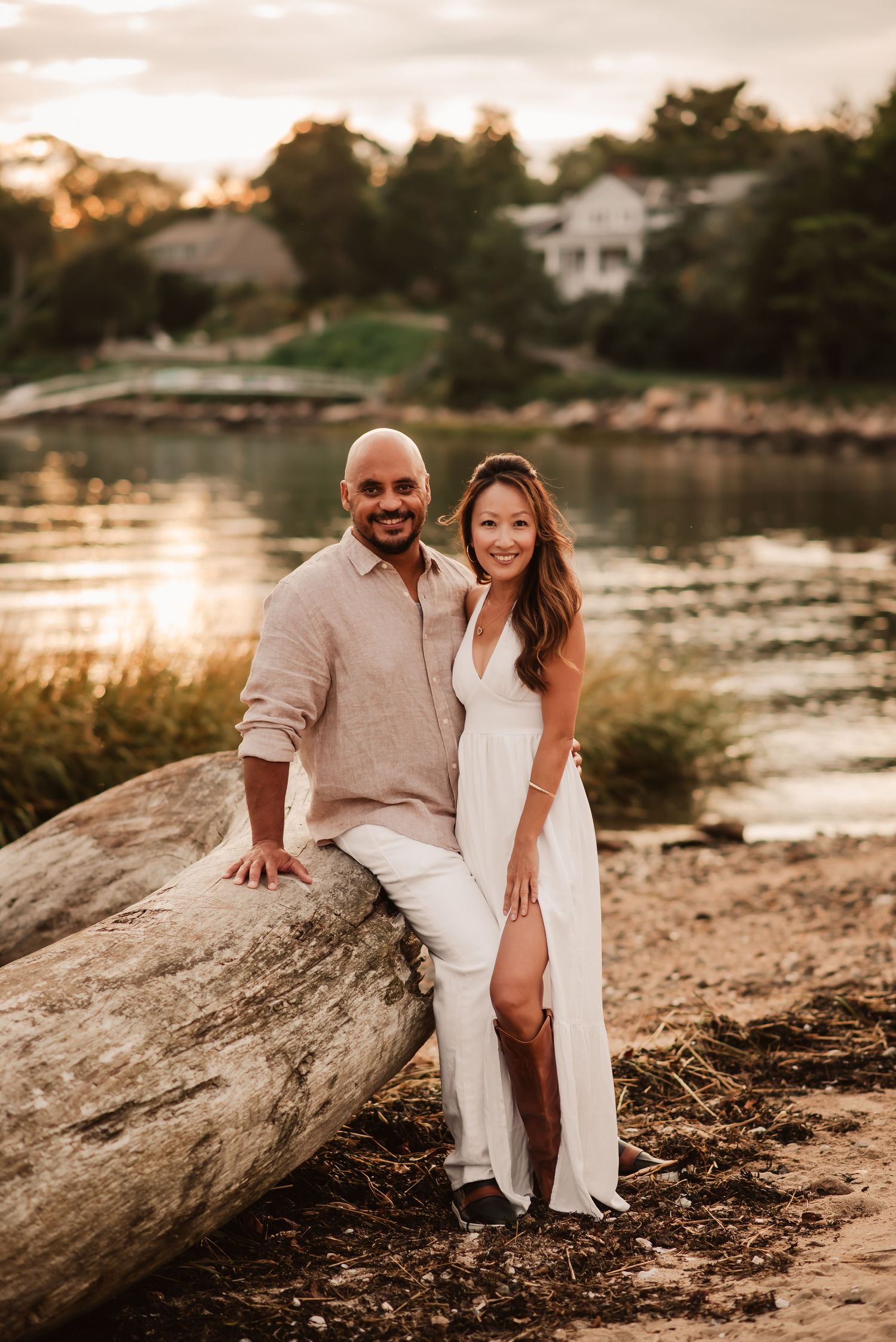 A couple in white and beige outfits sits together on a large rock by a lake during golden hour sunset.
