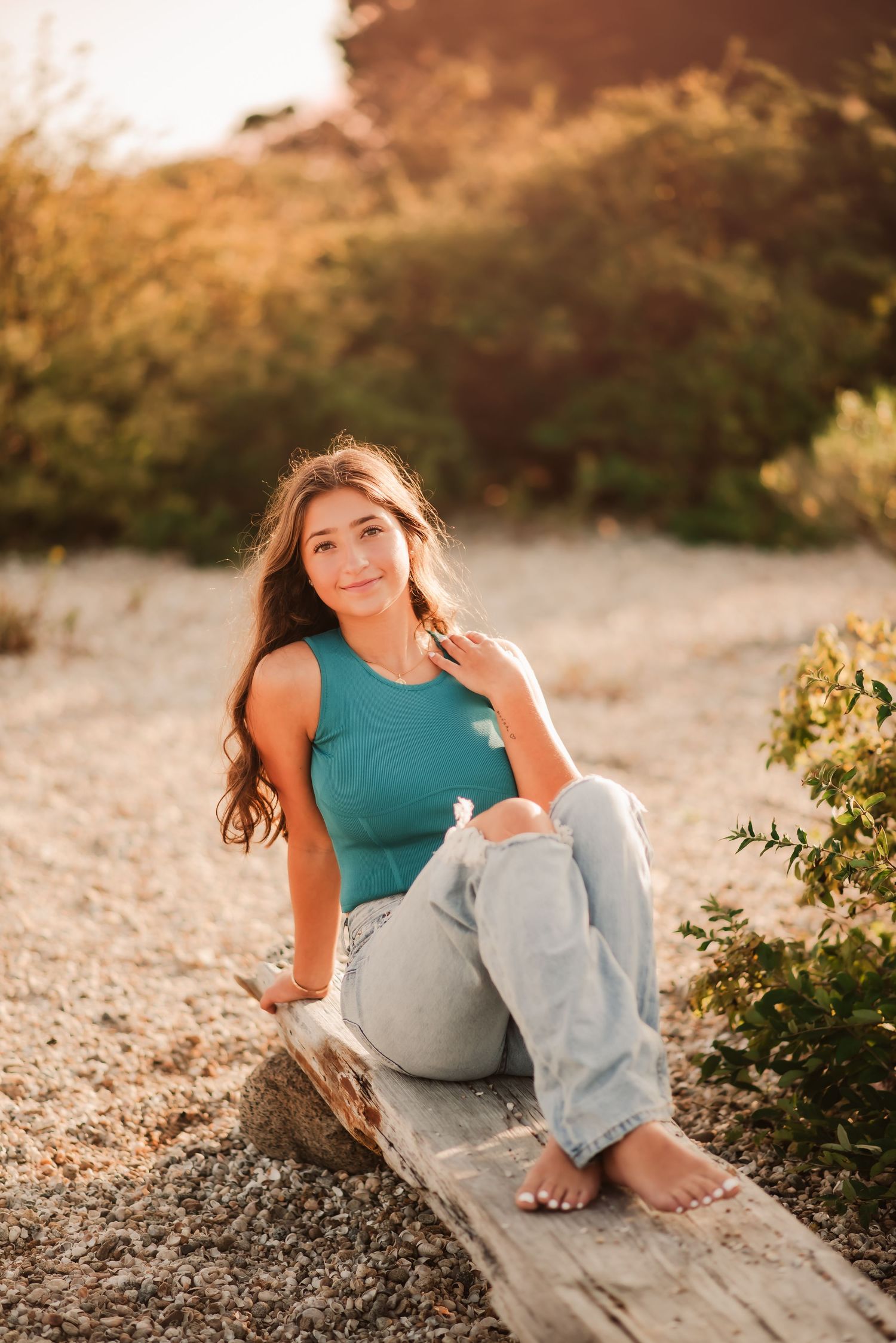 A person in a teal top and light jeans sits casually on a sandy beach during golden hour.