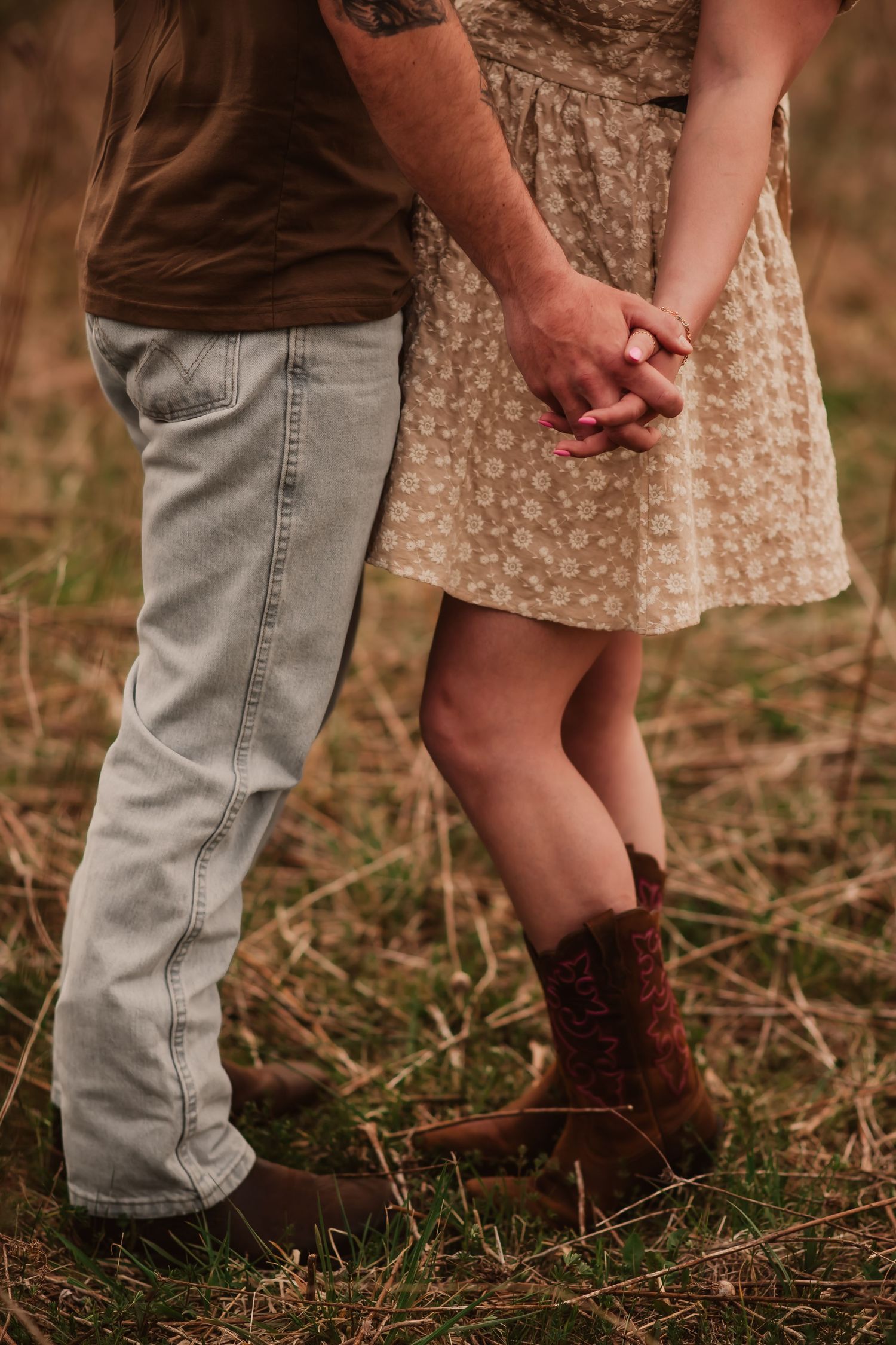 Close-up view of two people's legs and feet standing in brown grass wearing jeans and cowboy boots.