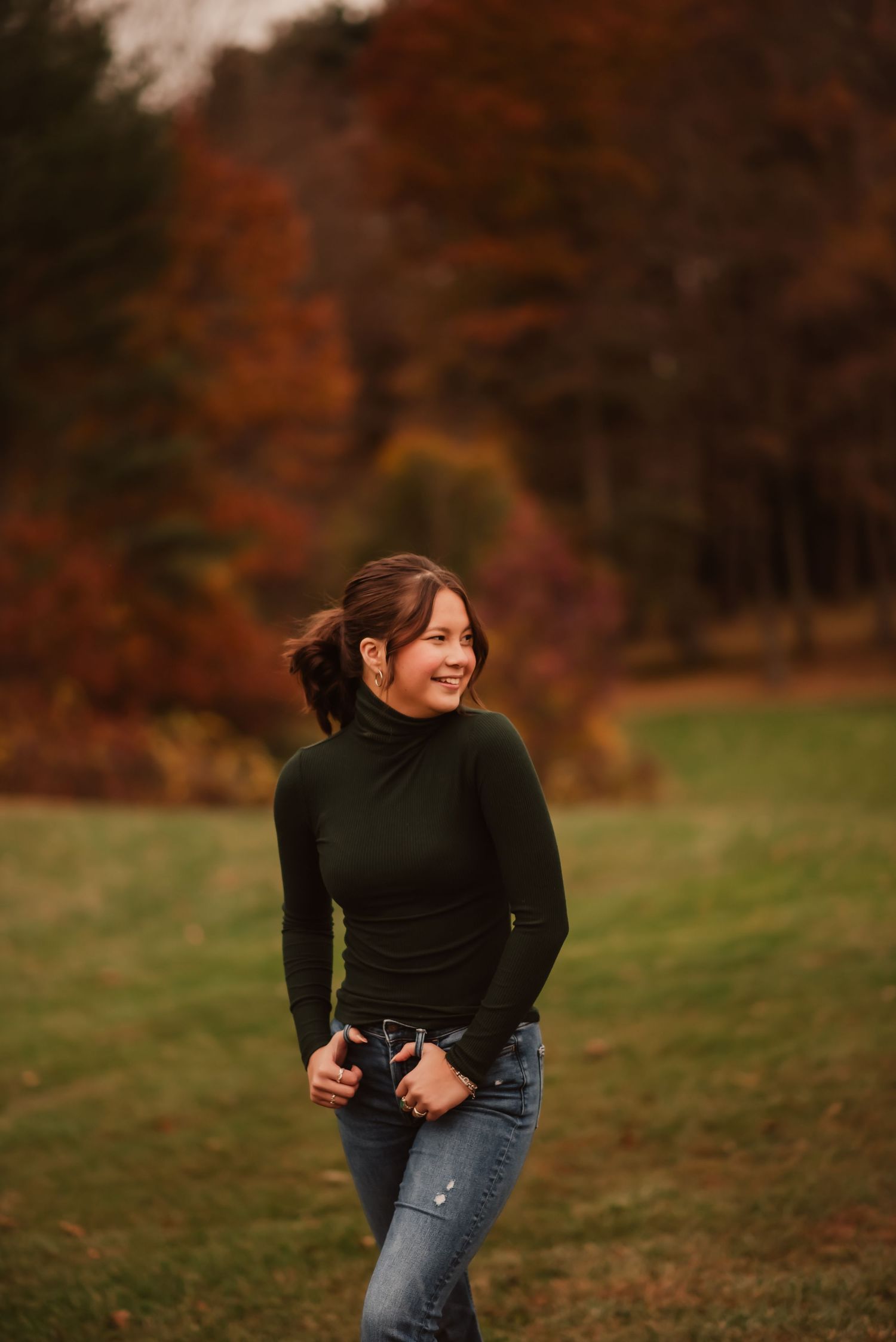 Person in black sweater and jeans laughs joyfully in autumn field with colorful fall foliage in background.