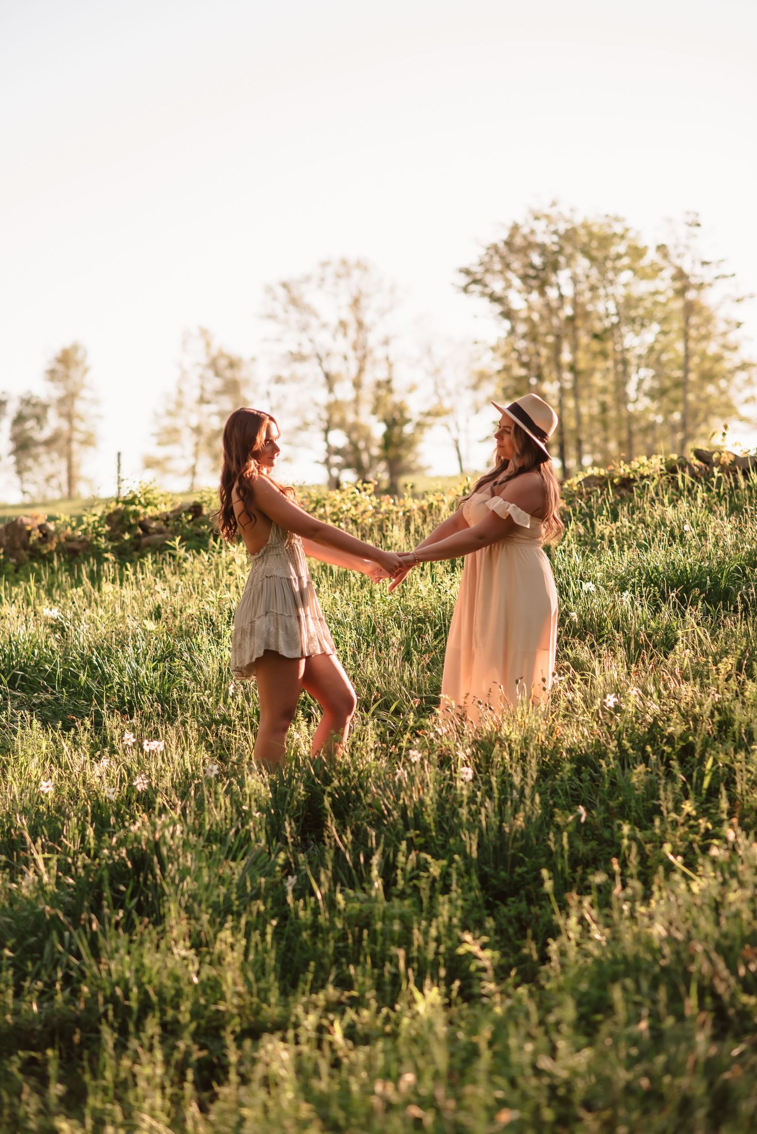 Two people in flowing dresses dance together in a sunlit meadow at golden hour among tall grass and wildflowers.