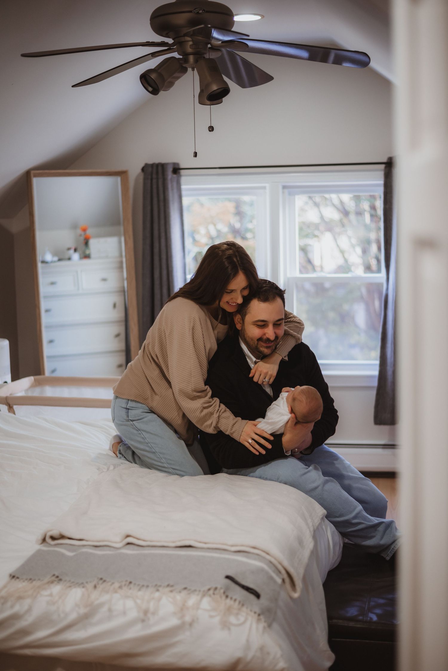 Couple sharing intimate moment with newborn baby on bed in cozy bedroom with ceiling fan and natural light.