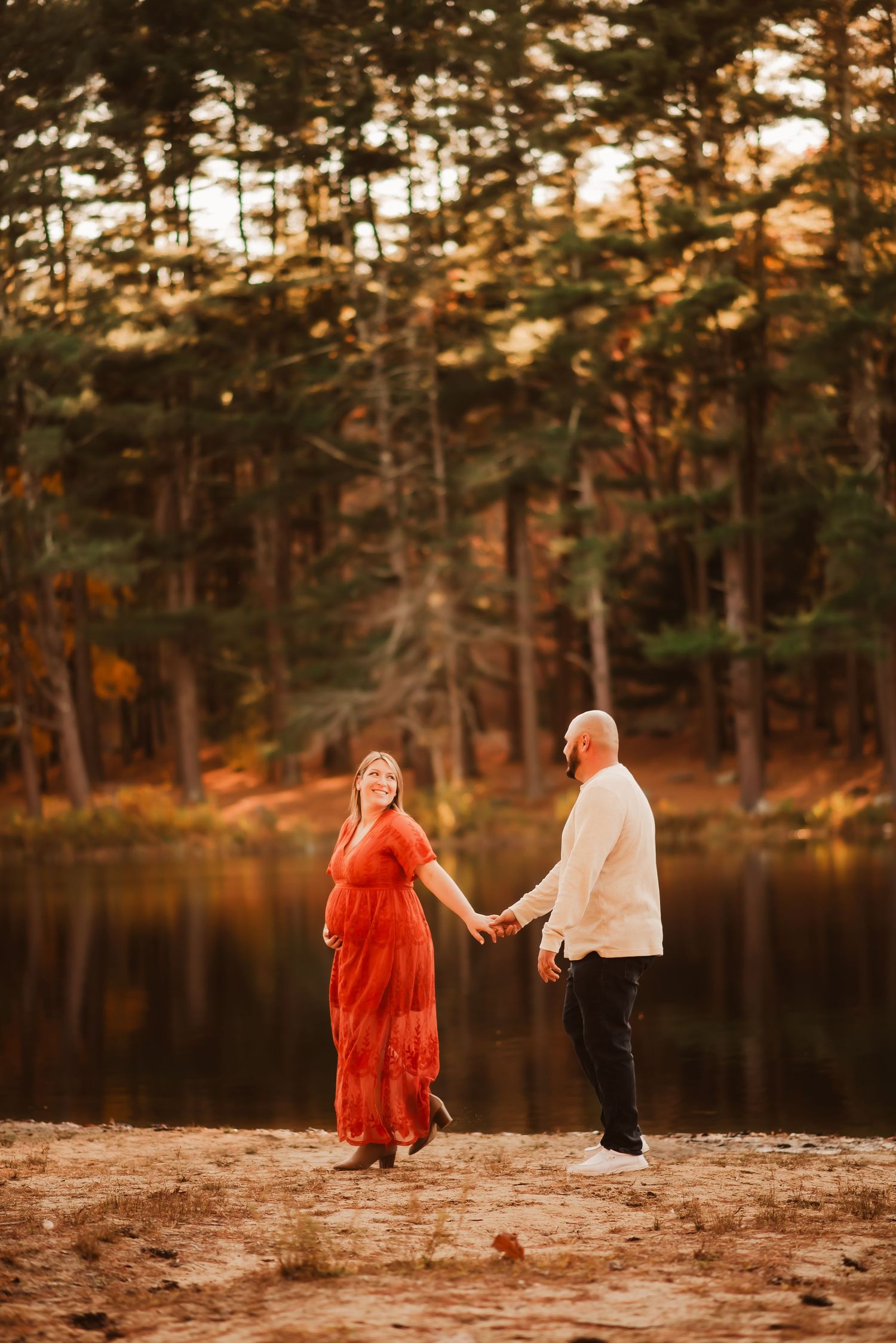 A couple walks hand in hand on a sandy beach at sunset with tall pine trees creating a warm autumn backdrop.