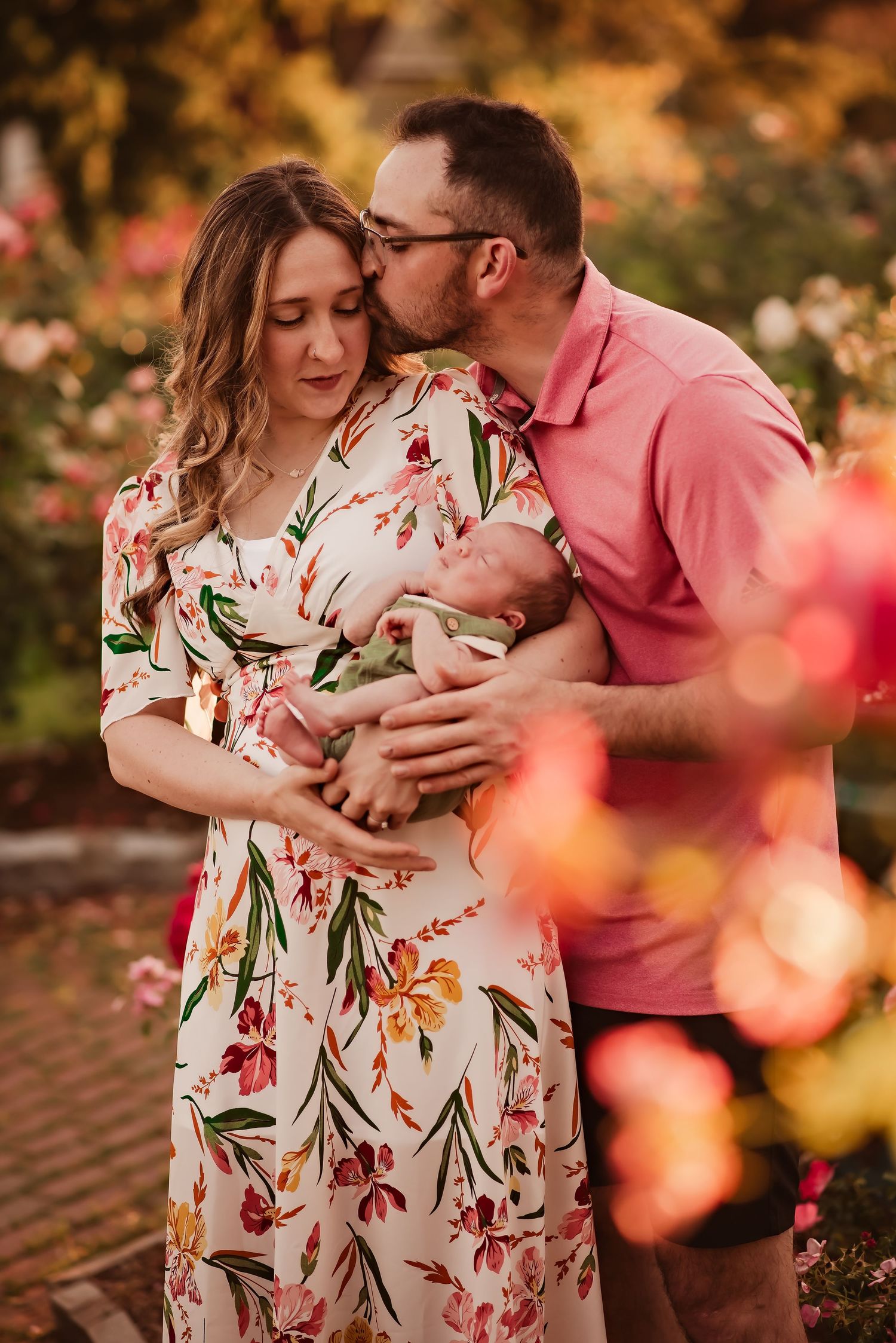 Romantic maternity photoshoot in a garden with floral dress and pink shirt as couple embraces amid autumn colors.