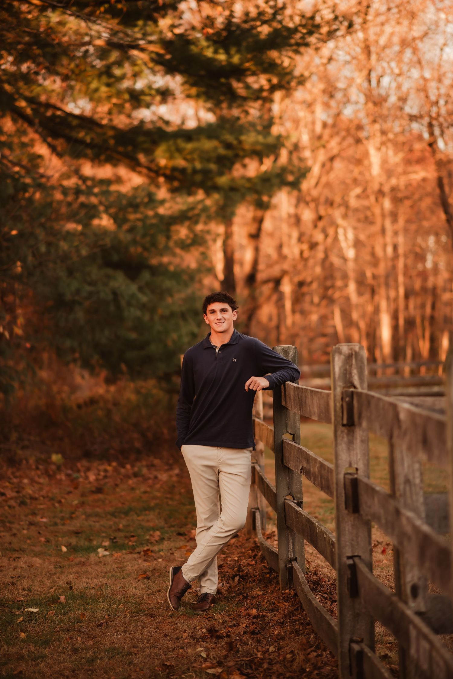 Person in casual attire leans against rustic wooden fence during golden autumn sunset in wooded area.