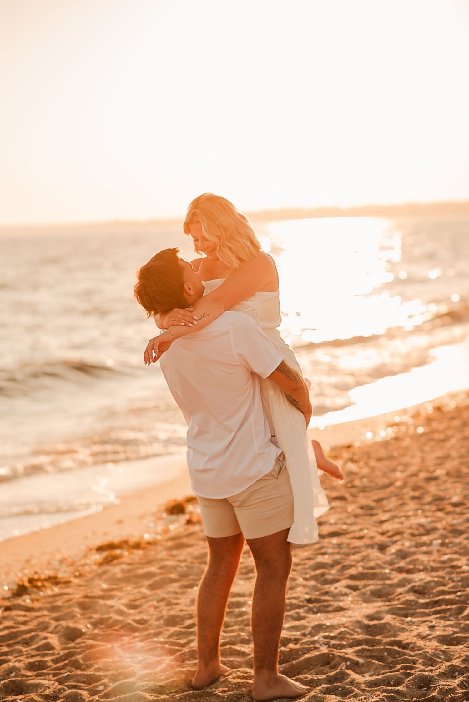 A romantic couple embraces on a sandy beach at sunset with golden sunlight and ocean waves in the background.