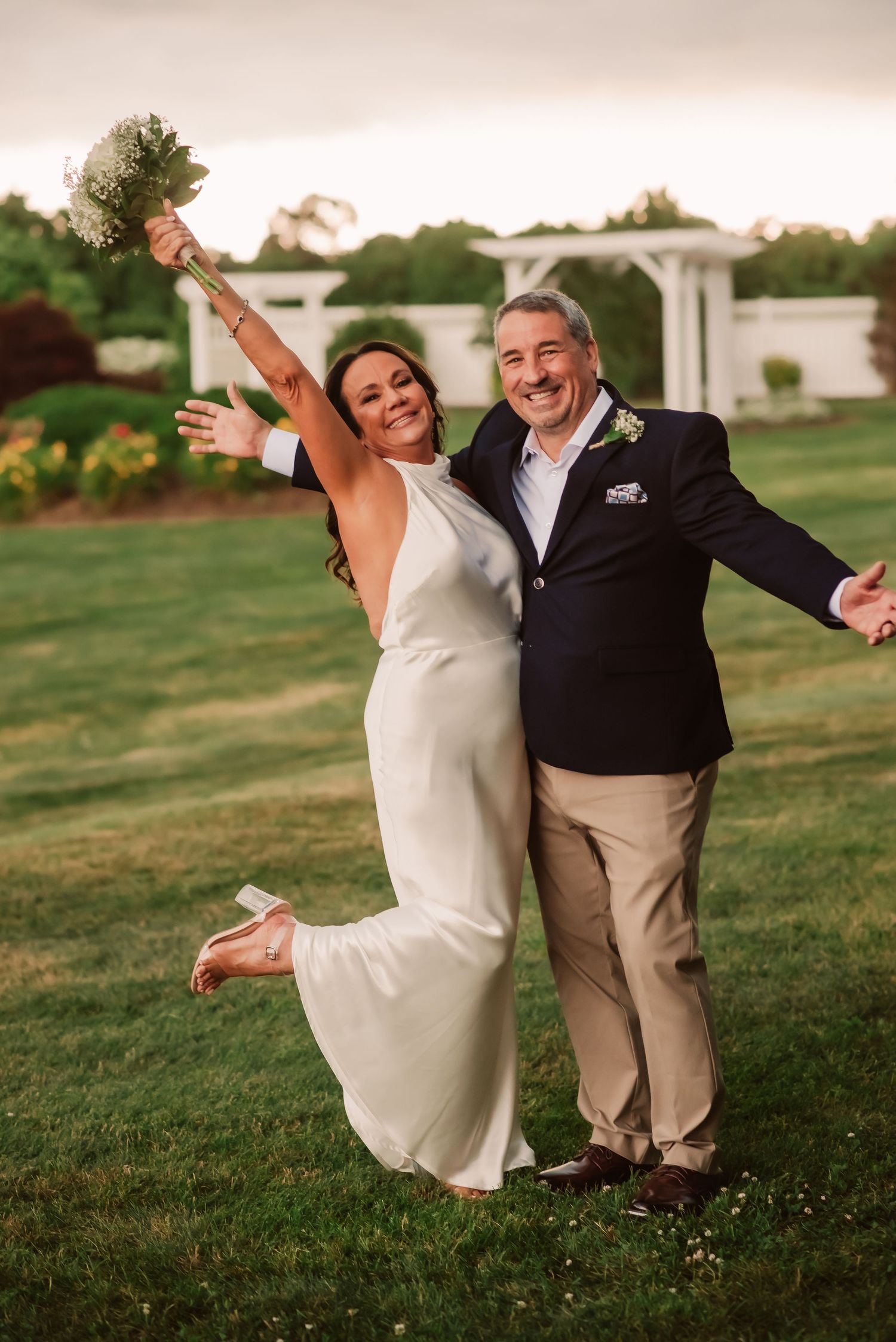 A joyful couple celebrates their wedding day on a green lawn with a white gazebo in the background.